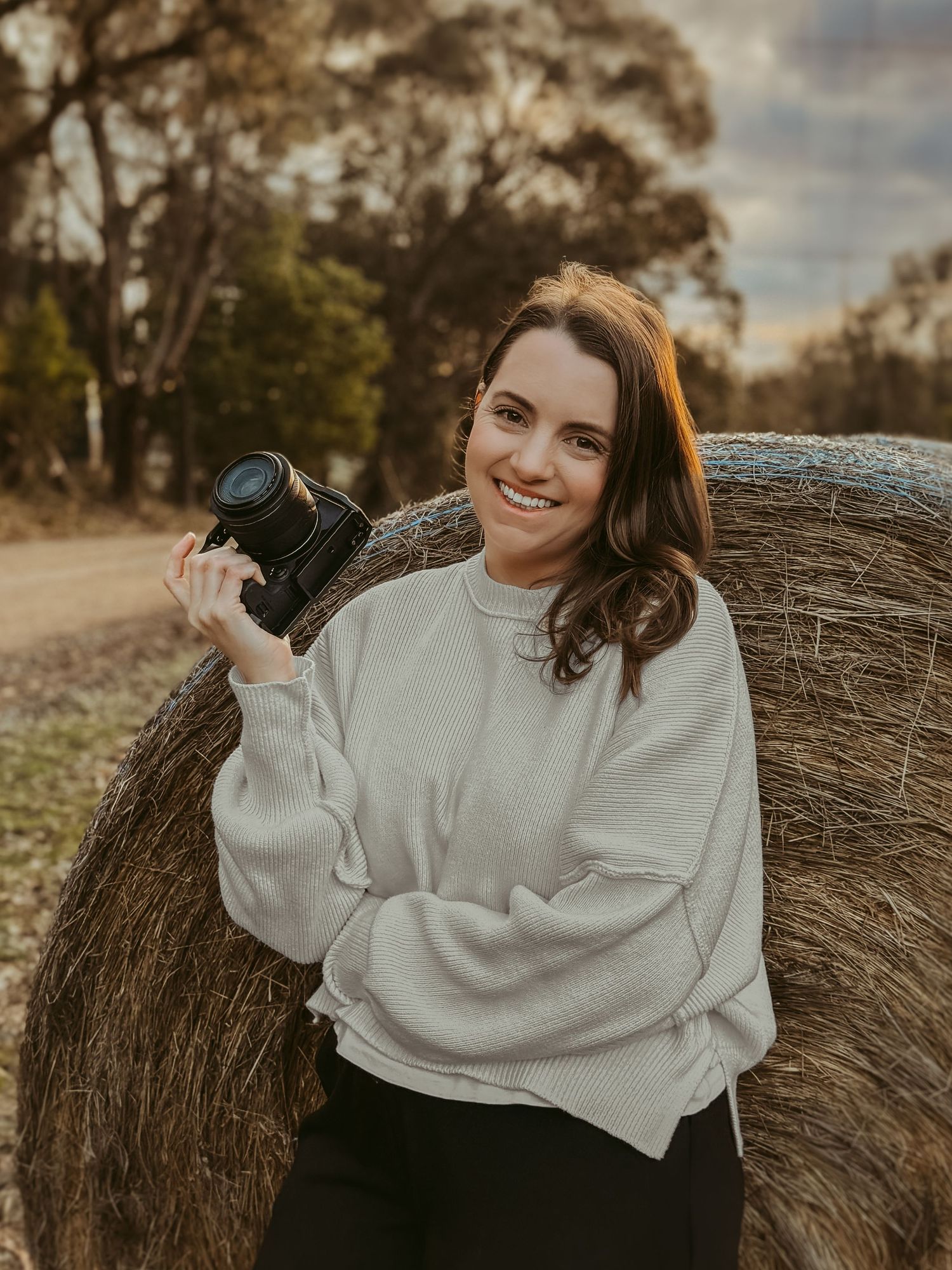 A person in a white sweatshirt posing with a camera near hay bales at sunset.