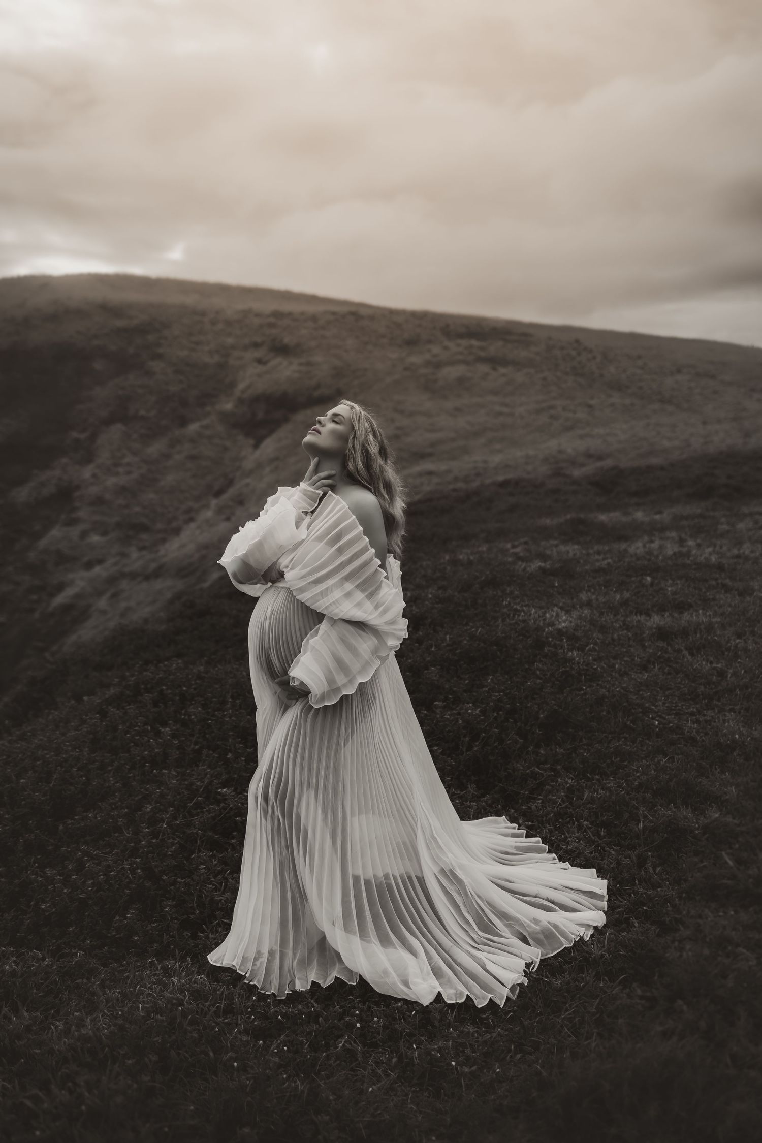 A dramatic black and white portrait features a pleated dress on a windswept hillside.