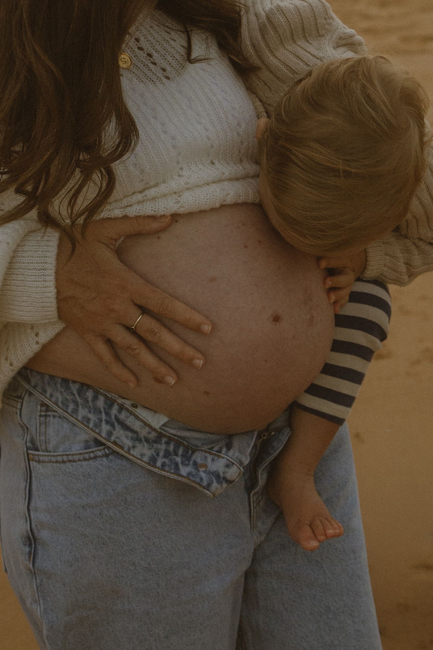 Pregnant woman in white sweater and jeans holding toddler against her bare baby bump outdoors.