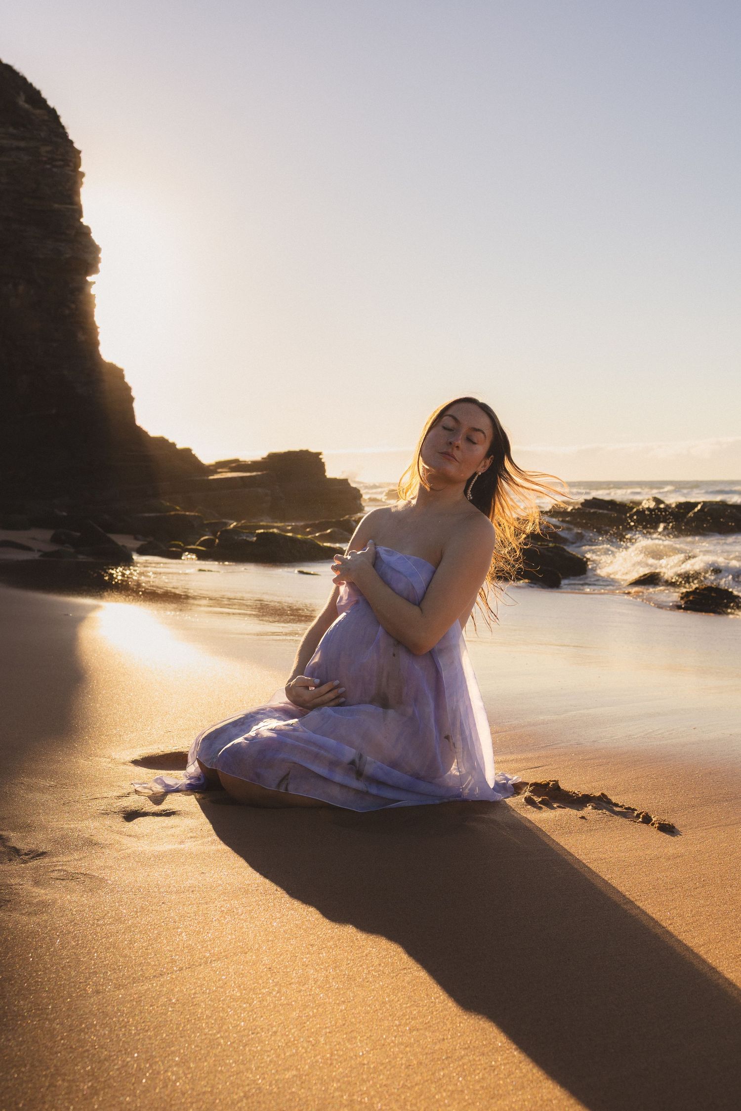Pregnant woman in lavender dress kneels on sandy beach cradling her belly during golden hour sunset.