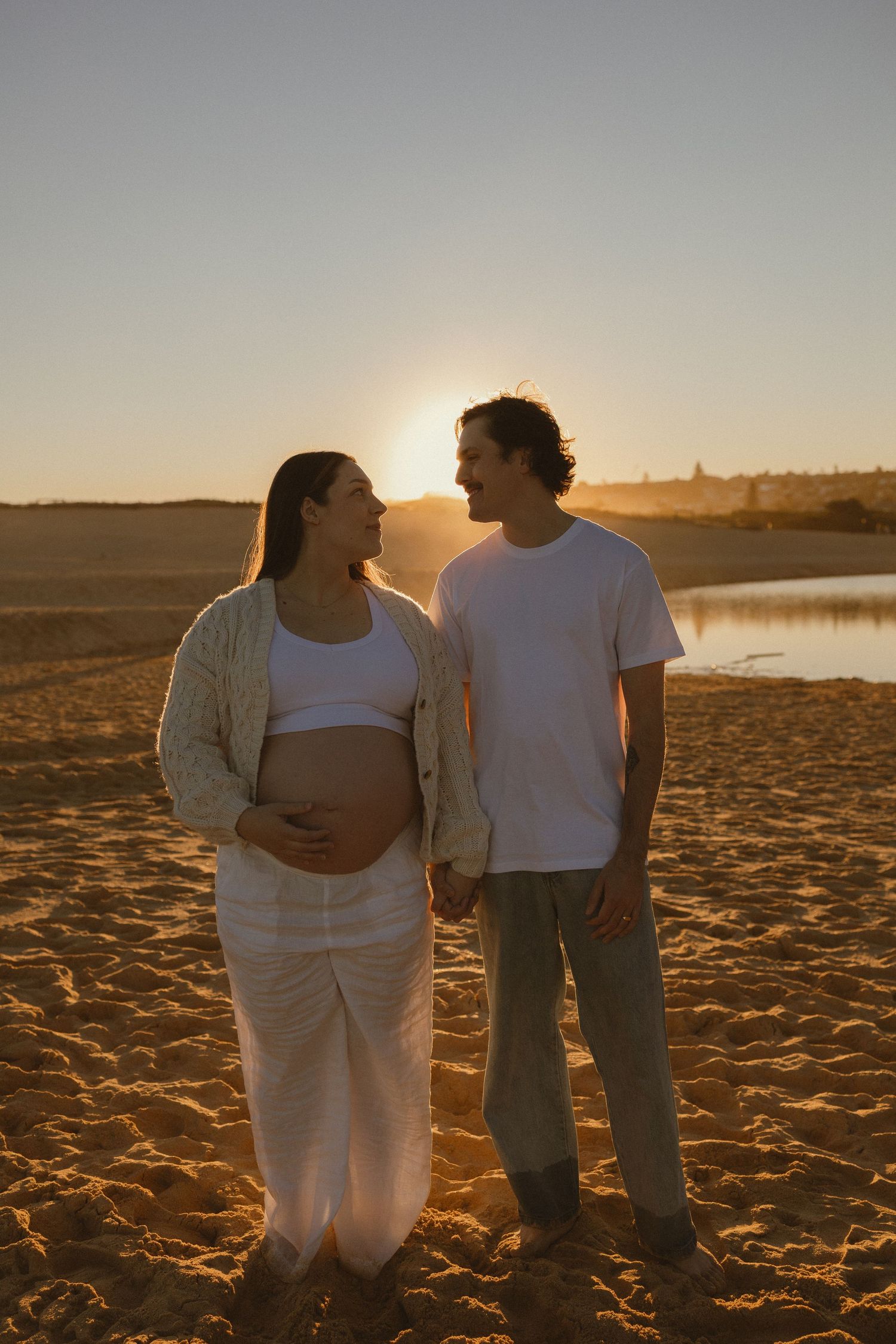 Pregnant couple holding hands on sandy beach at golden sunset, woman cradling baby bump in white outfit.