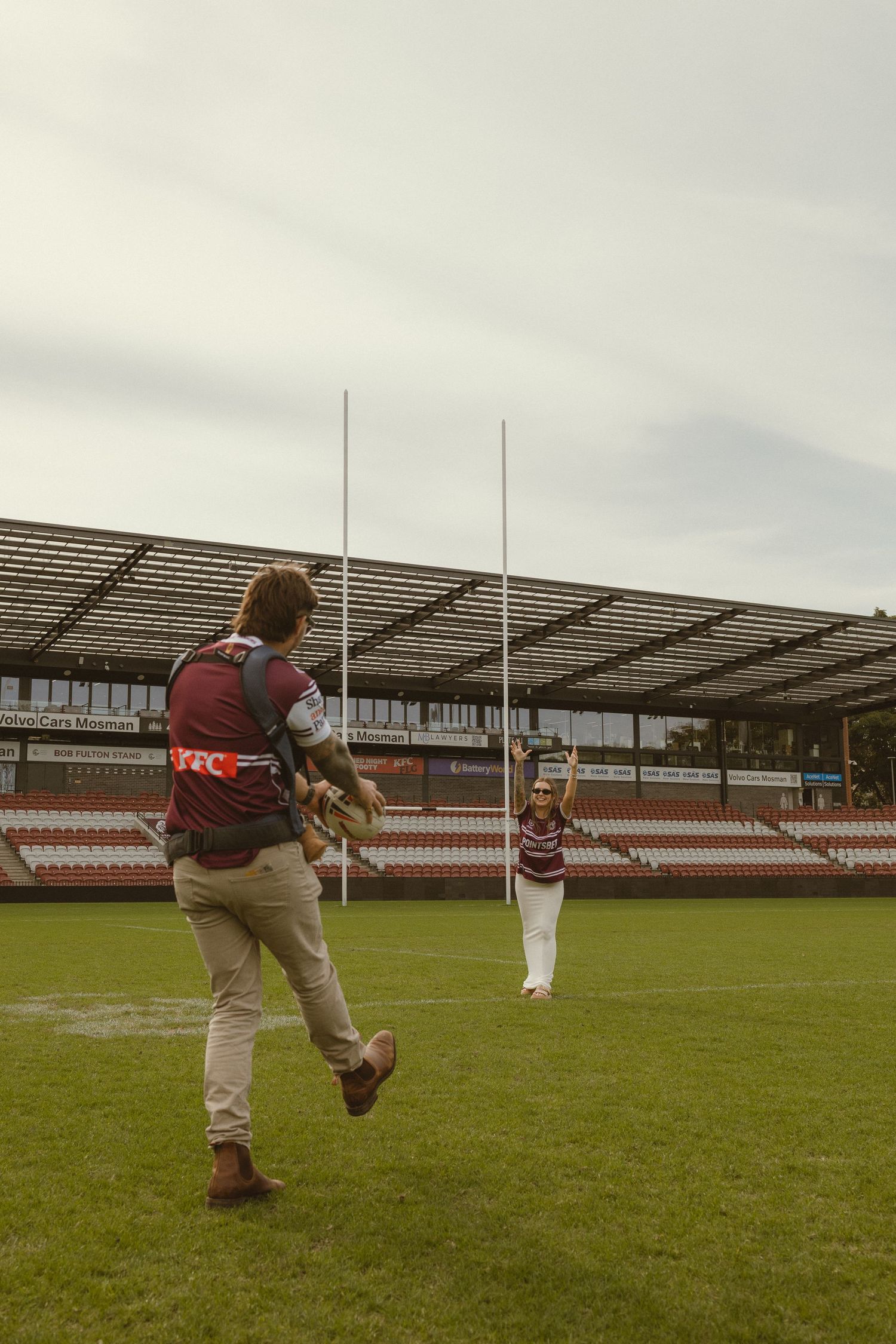 Two rugby players practicing kicks on a green field at an empty stadium under an overcast sky.