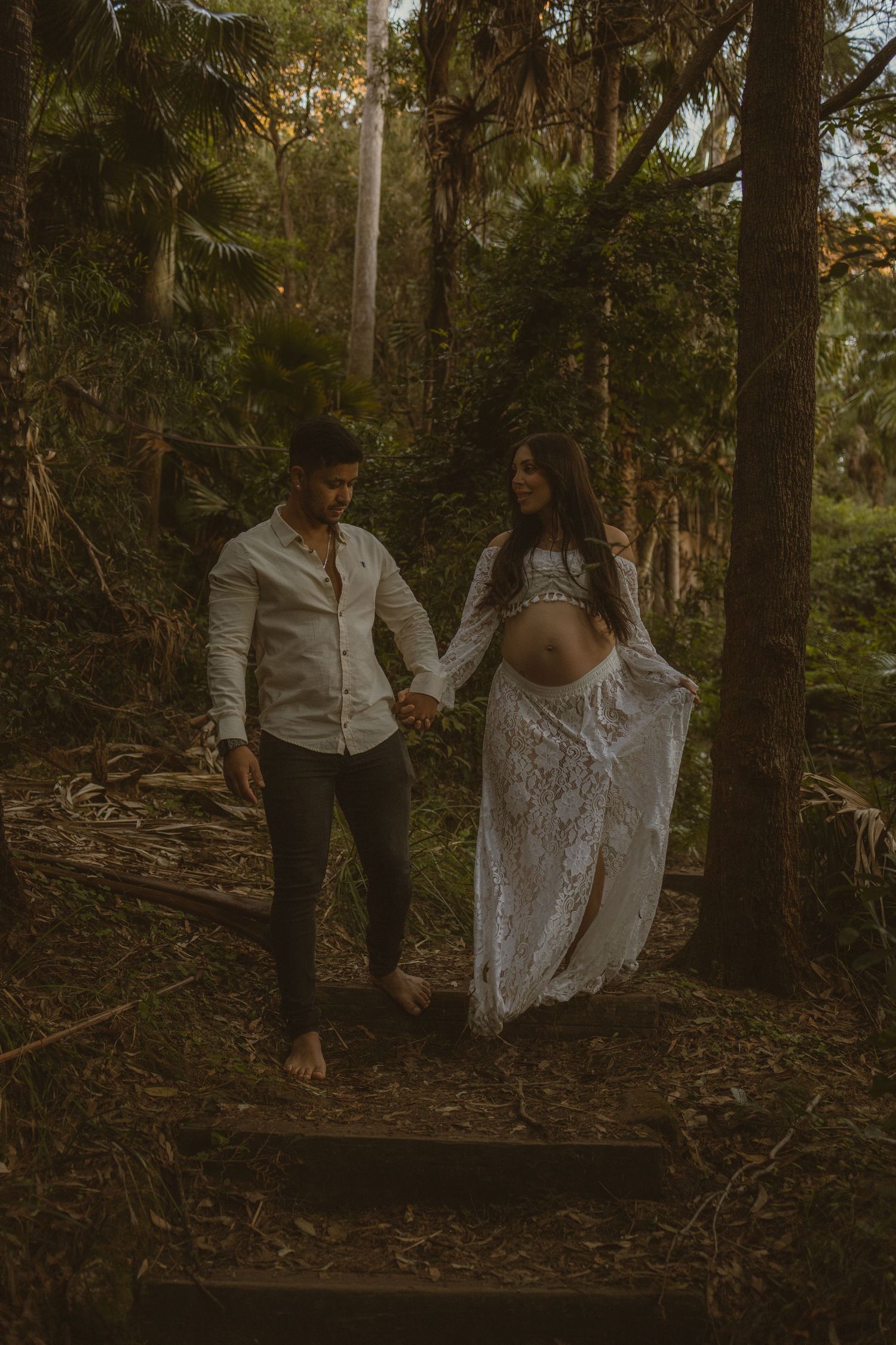 Pregnant couple holding hands walking through lush forest, woman in white lace maternity gown, man in white shirt and dark pants.