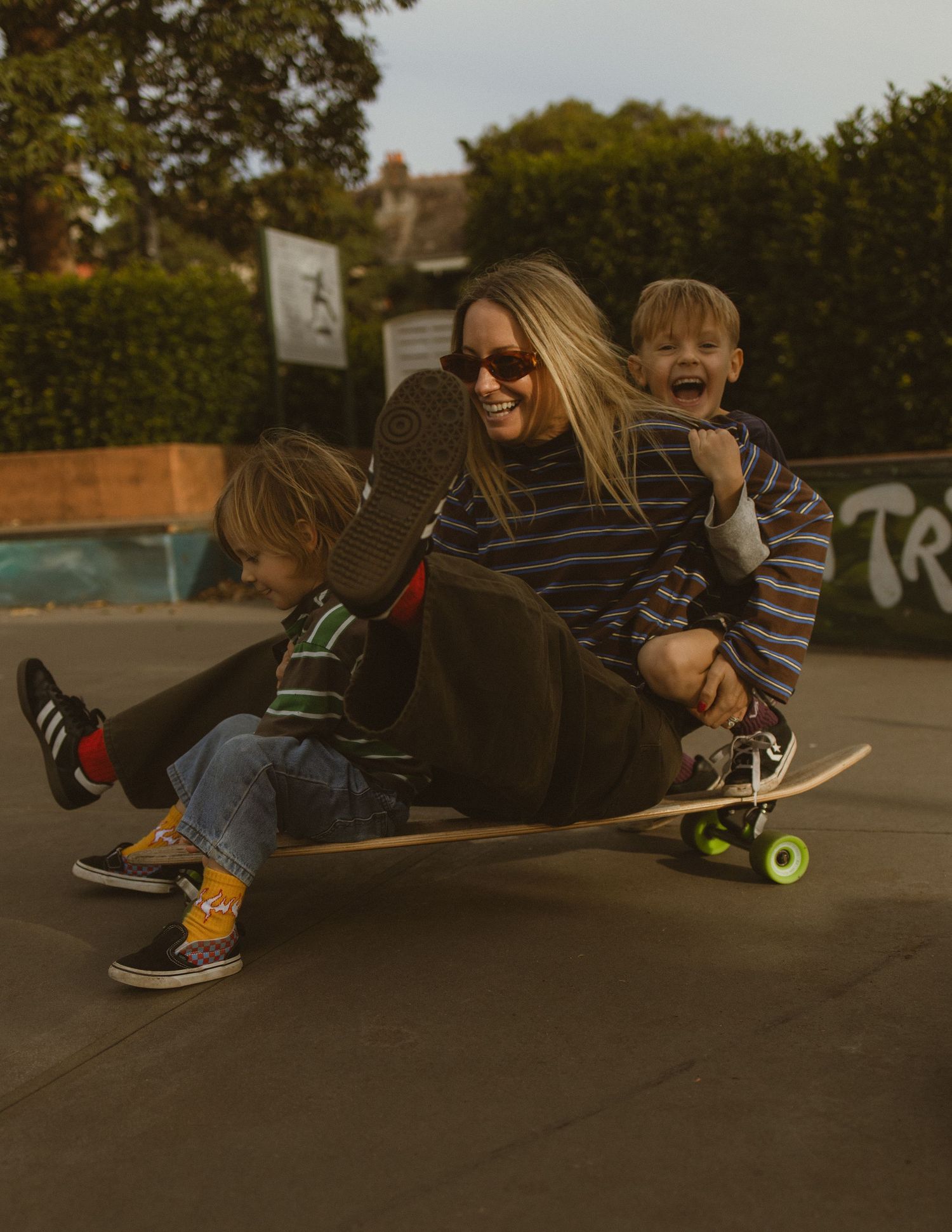 A woman and two young children laugh while sitting together on a skateboard at an outdoor skate park.