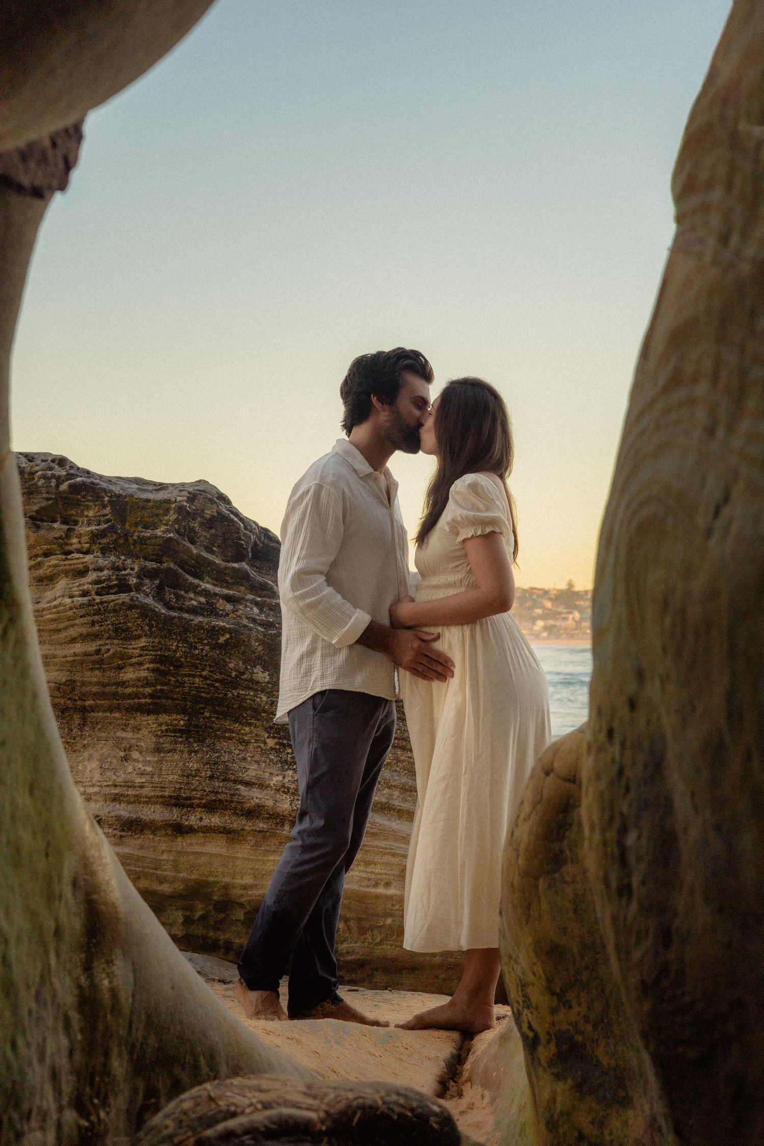 Couple sharing a romantic kiss on rocky beach at sunset, woman in white dress with visible baby bump.