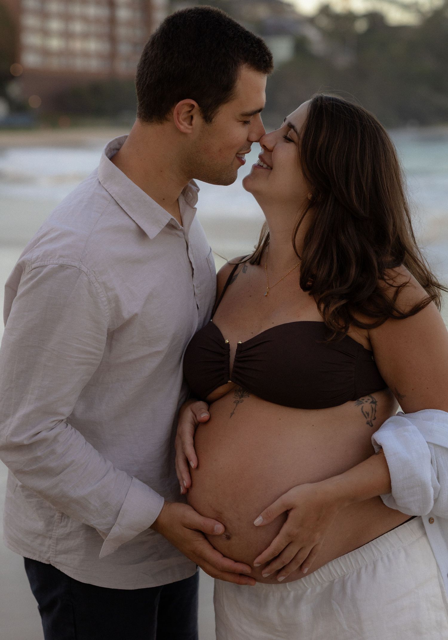 A romantic maternity photoshoot at sunset shows a couple embracing by the water in matching white outfits.