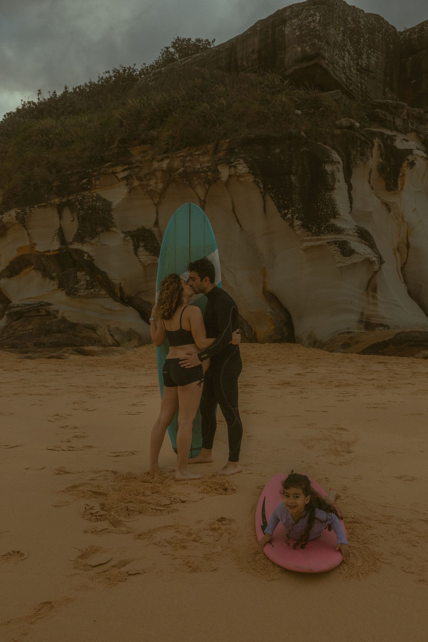 Couple kissing on sandy beach holding blue surfboard while young girl plays on pink bodyboard near rocky cliffs.