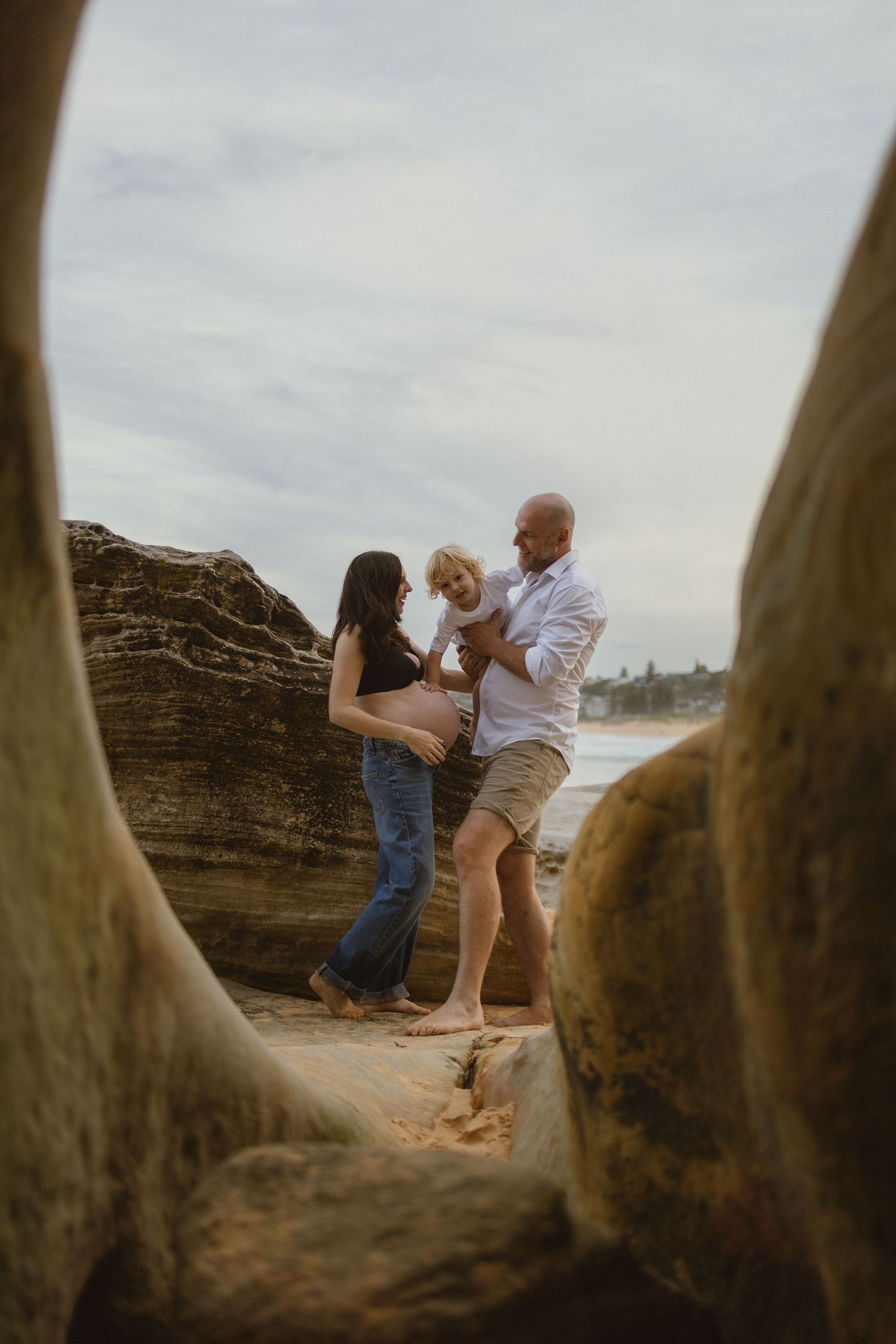Pregnant woman and bald man holding toddler on sandy beach framed by rocks, maternity family photo.