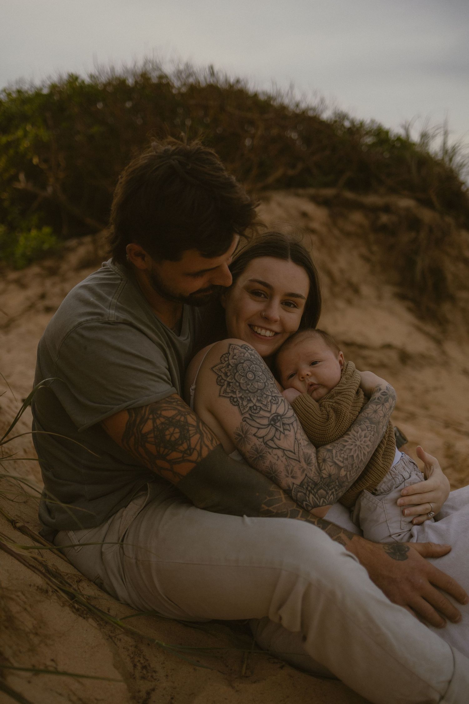 A couple with tattoos sits on sandy dunes, tenderly holding their newborn baby in a warm, intimate family portrait.