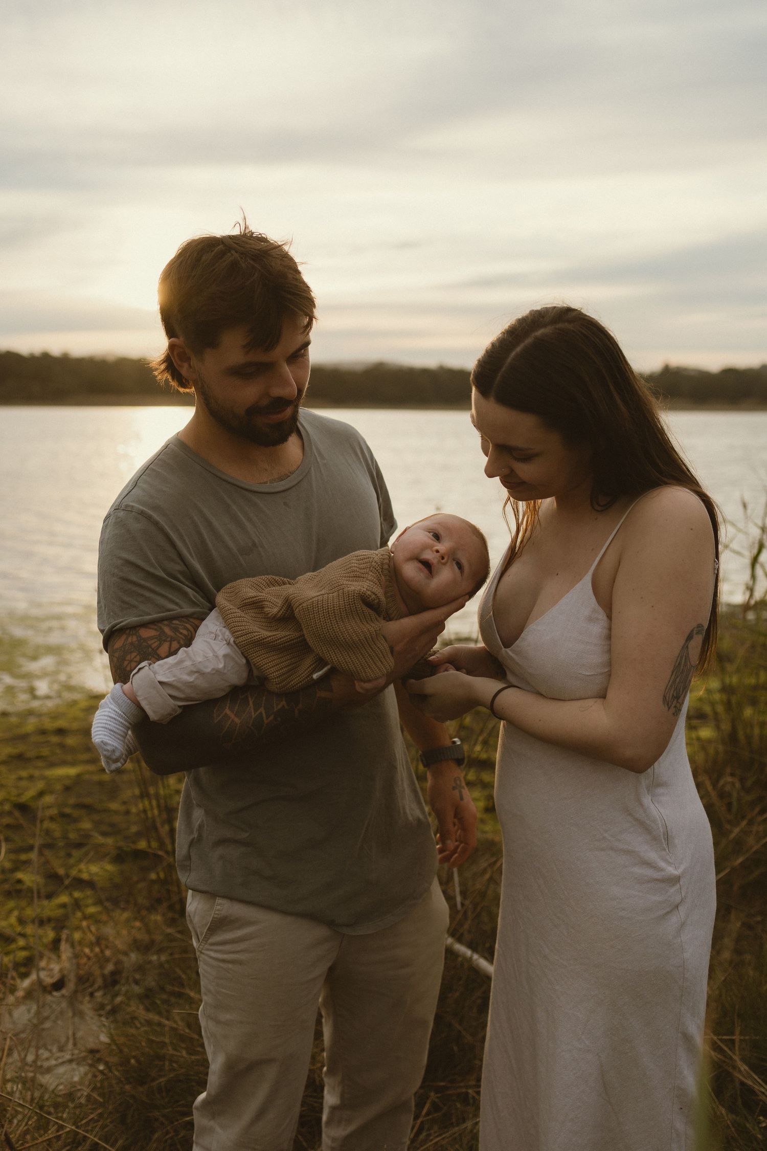 Young couple holding their newborn baby outdoors near a lake at golden hour sunset.