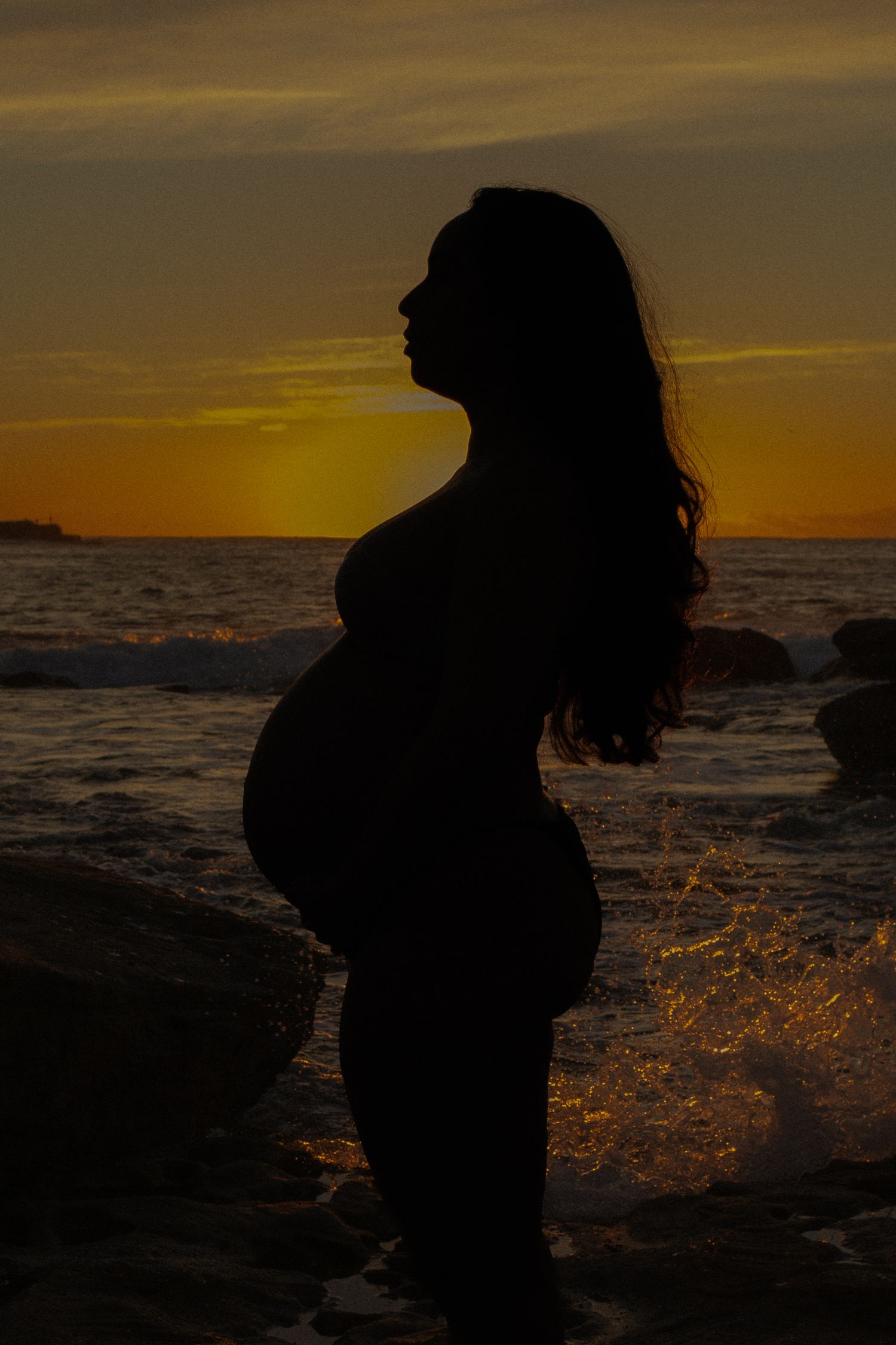 Silhouette of pregnant woman standing on rocky beach at golden sunset with ocean waves in background.
