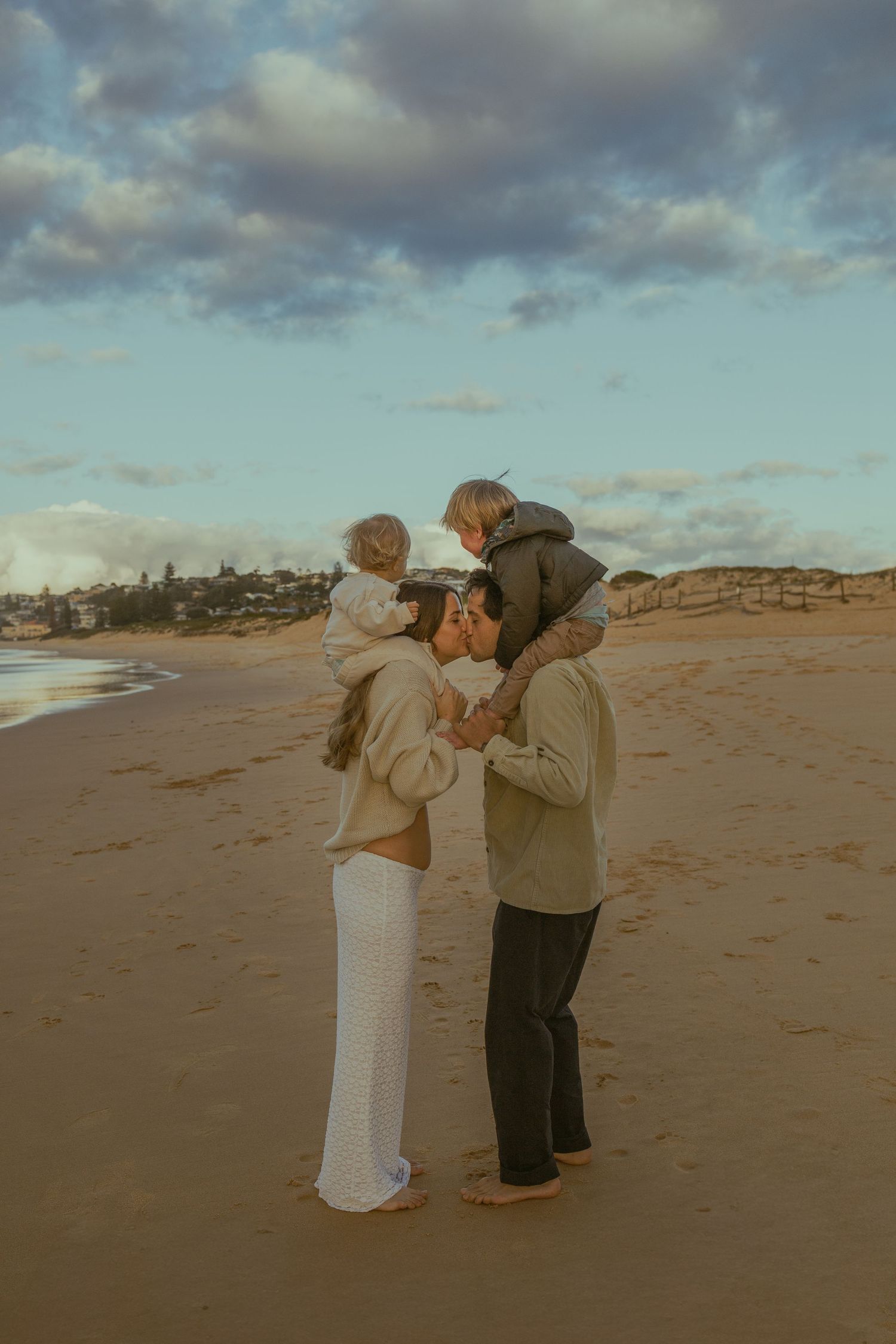 Family of four on sandy beach at sunset, parents holding toddlers on shoulders, ocean waves in background.