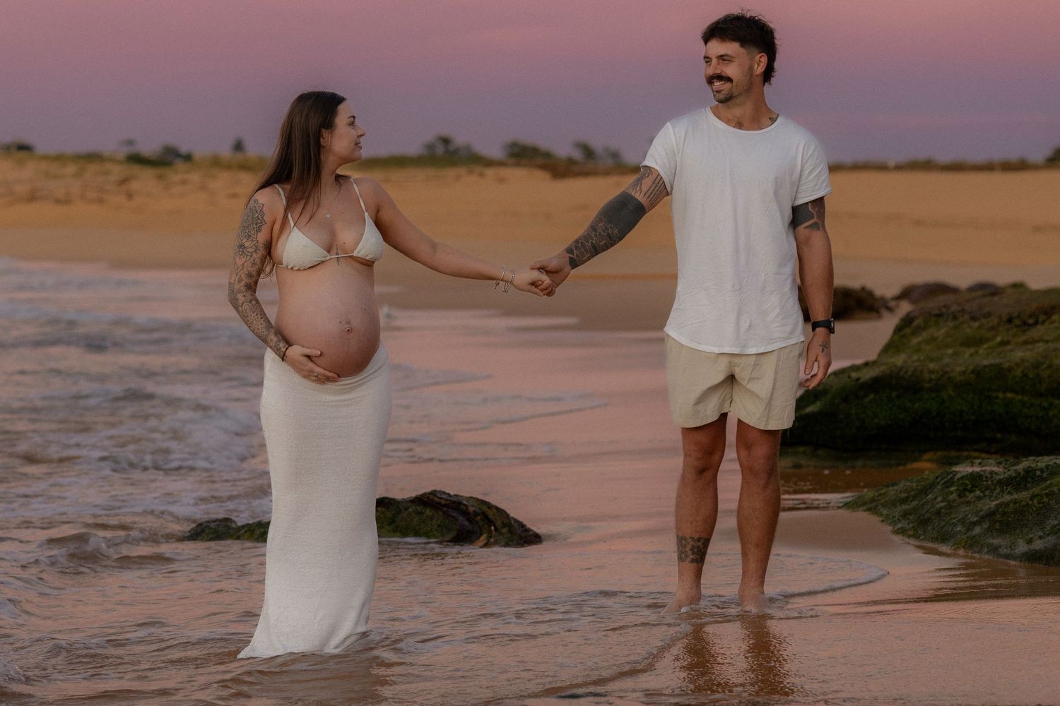 Pregnant woman in white dress holds hands with tattooed man on beach at sunset during maternity photoshoot.