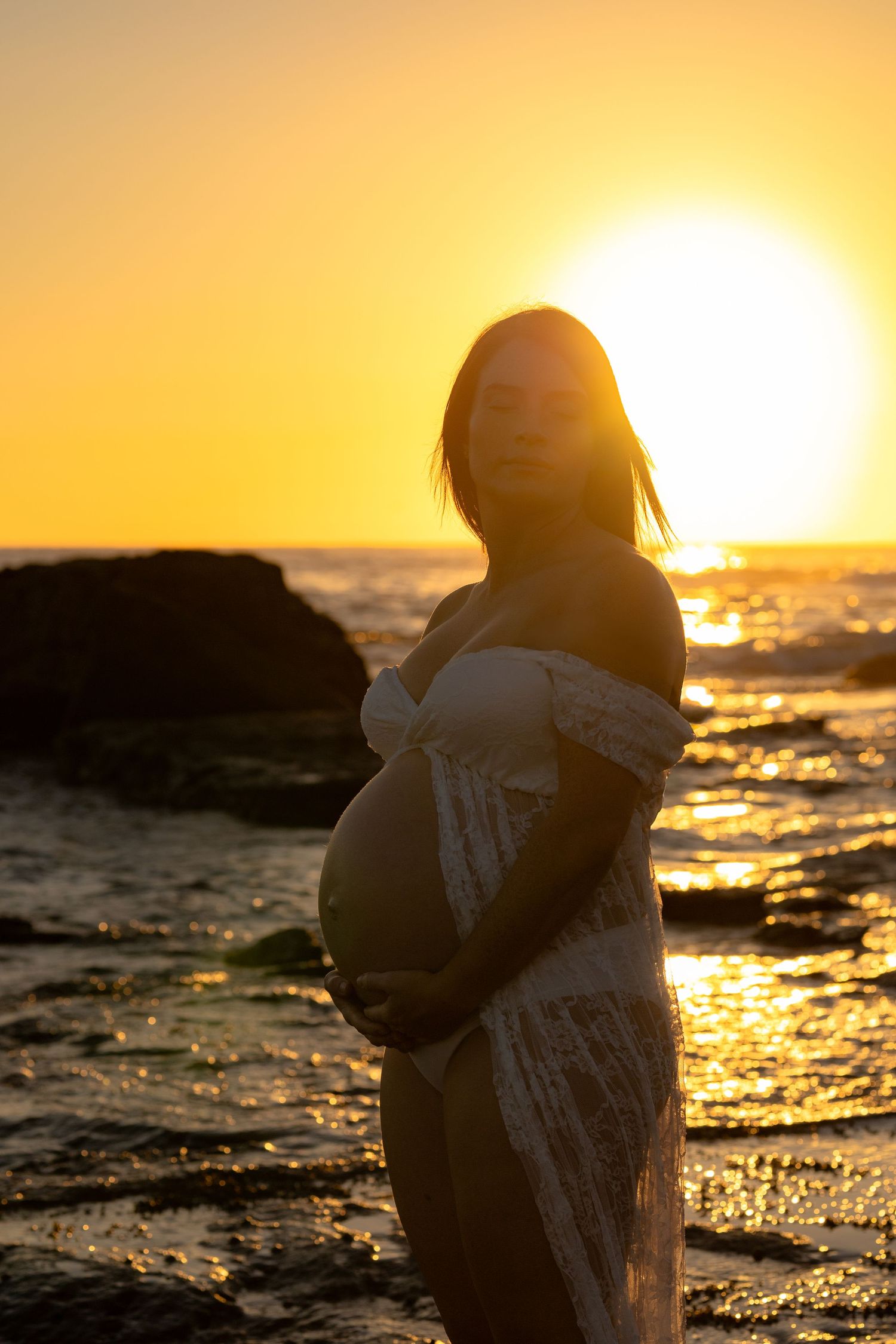 Pregnant woman in white lace dress holding belly at golden sunset on rocky beach.