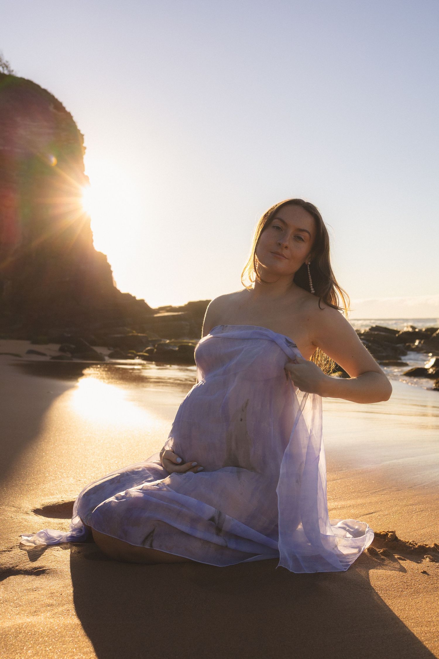 Pregnant woman wrapped in soft blue fabric sits on sunlit beach with rocky cliffs and ocean backdrop.