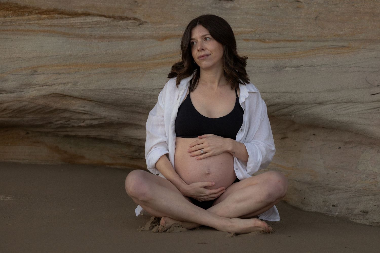 Pregnant woman sitting cross-legged on sandy beach, wearing black bikini top and open white shirt, cradling her belly.
