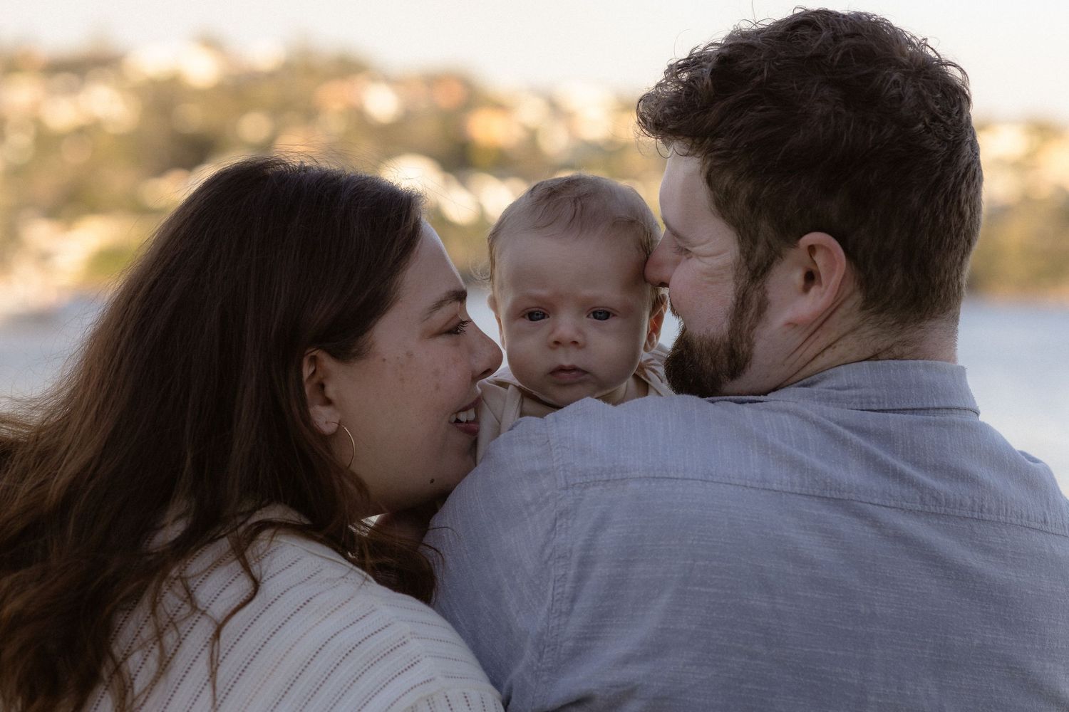 A couple lovingly kisses their baby outdoors at golden hour in a warm family portrait.