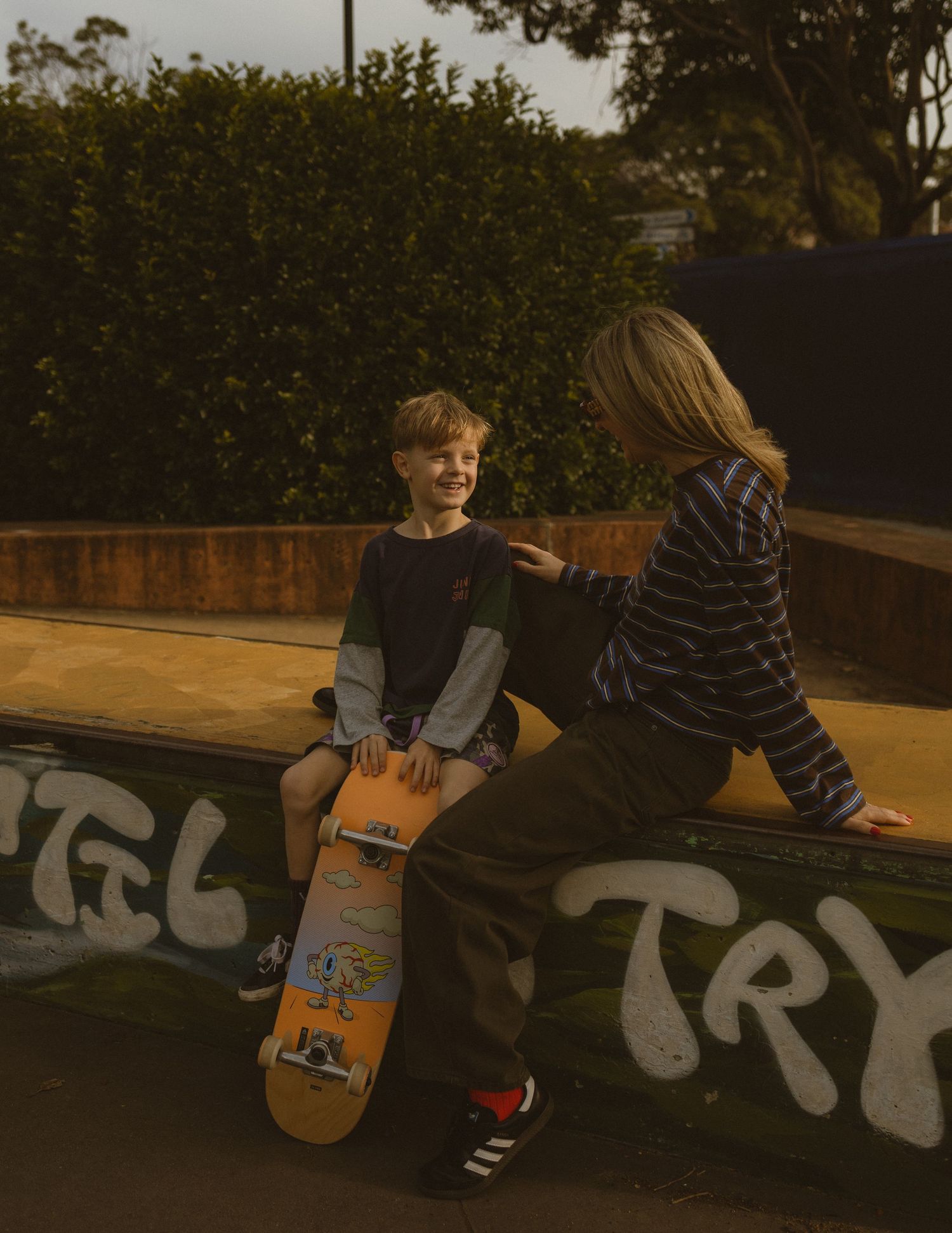 Two kids at a skate park, one holding an orange skateboard, sitting on a graffiti wall surrounded by green hedges.