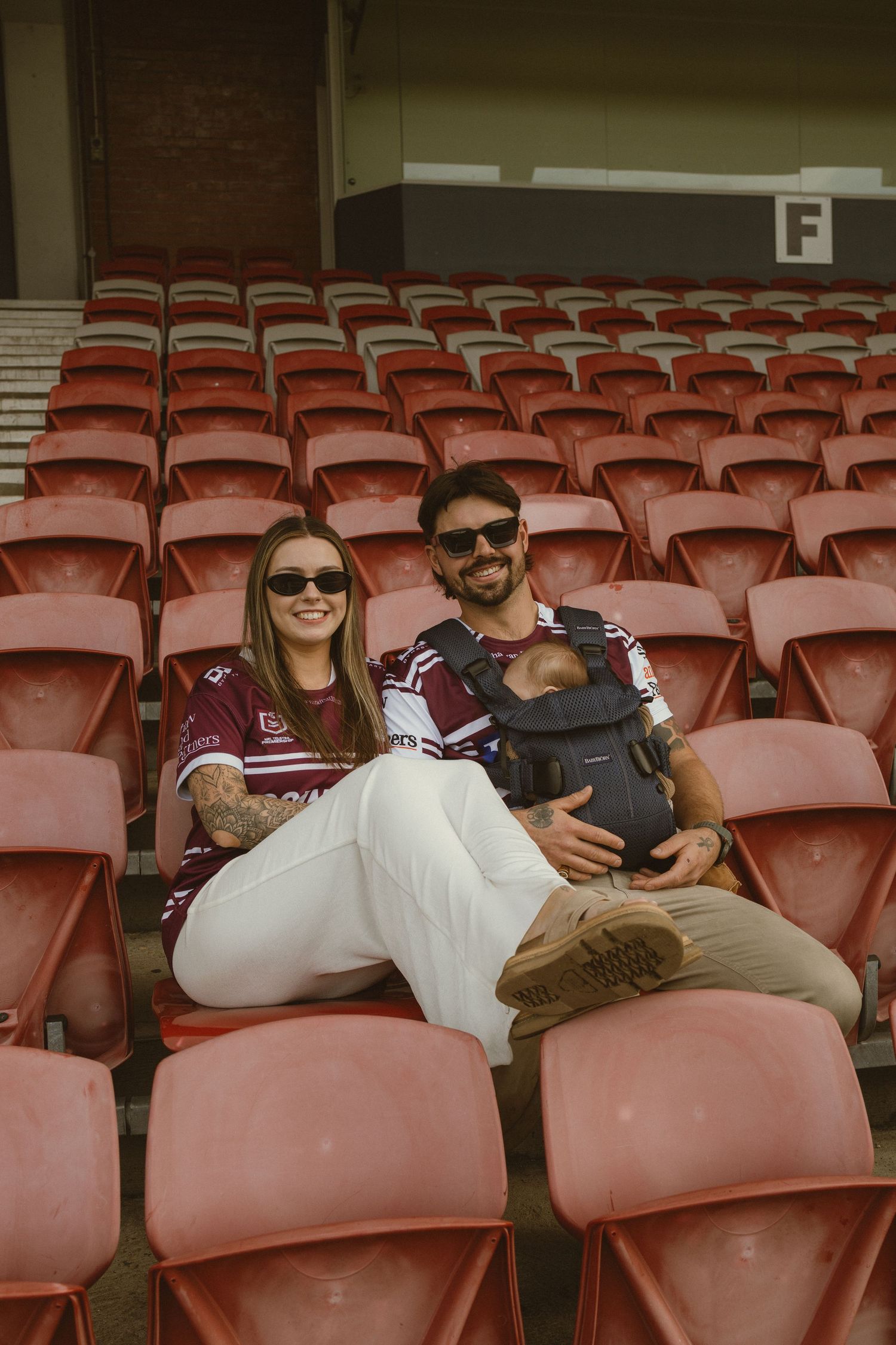 Couple in matching maroon jerseys sitting in red stadium seats, man holding baby in carrier, smiling at camera.