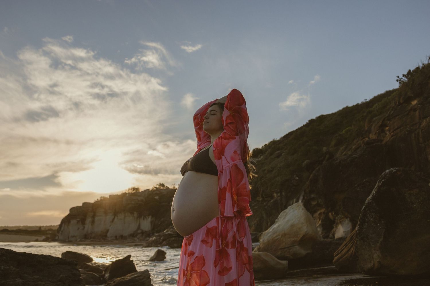 Pregnant woman in pink floral dress poses on rocky beach at sunset with cliffs in background.