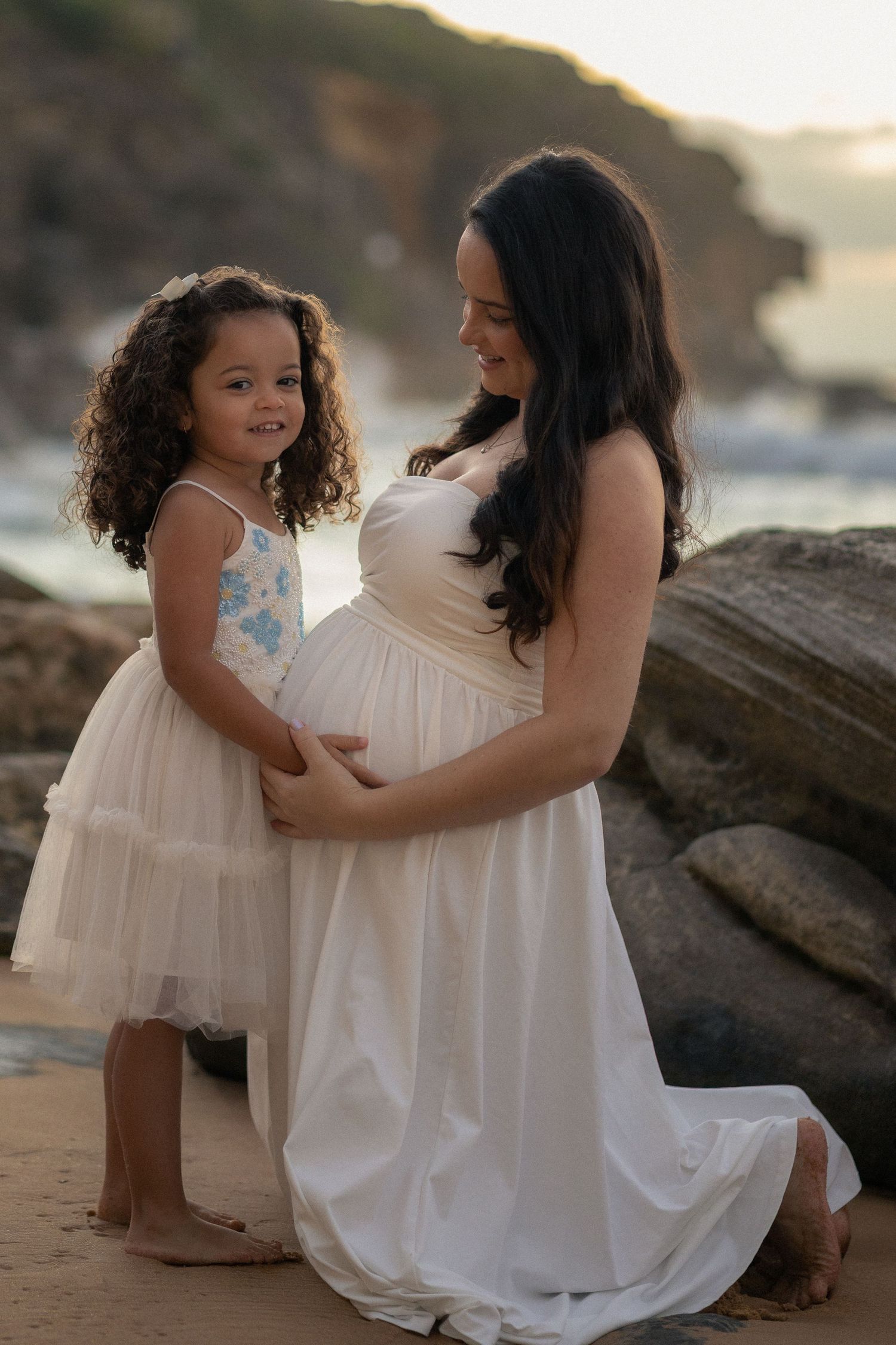 A mother and child in white dresses share a tender moment on the beach at sunset.