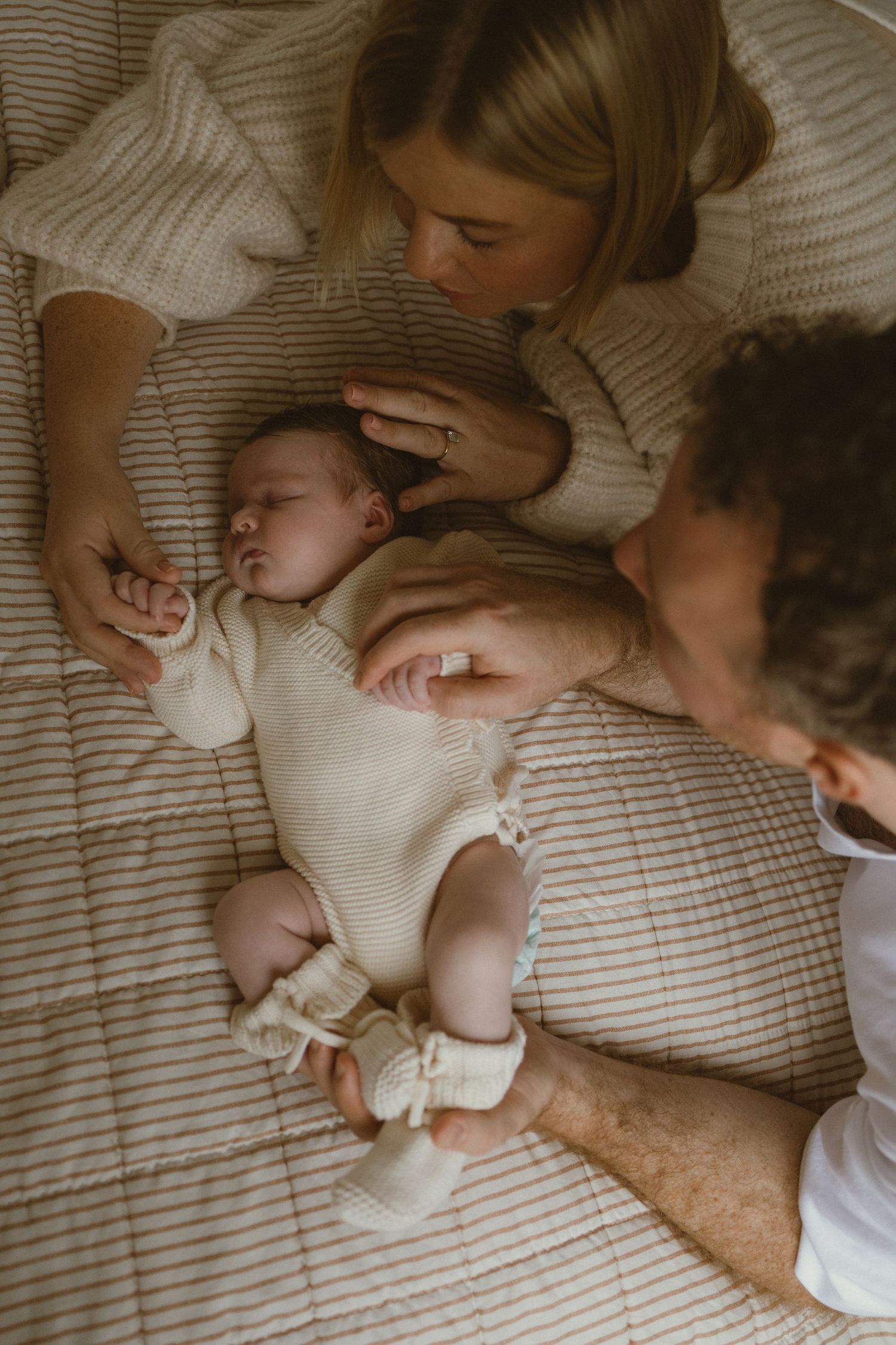 Parents lovingly gazing at their sleeping newborn baby dressed in a cream knit outfit on a striped bed.