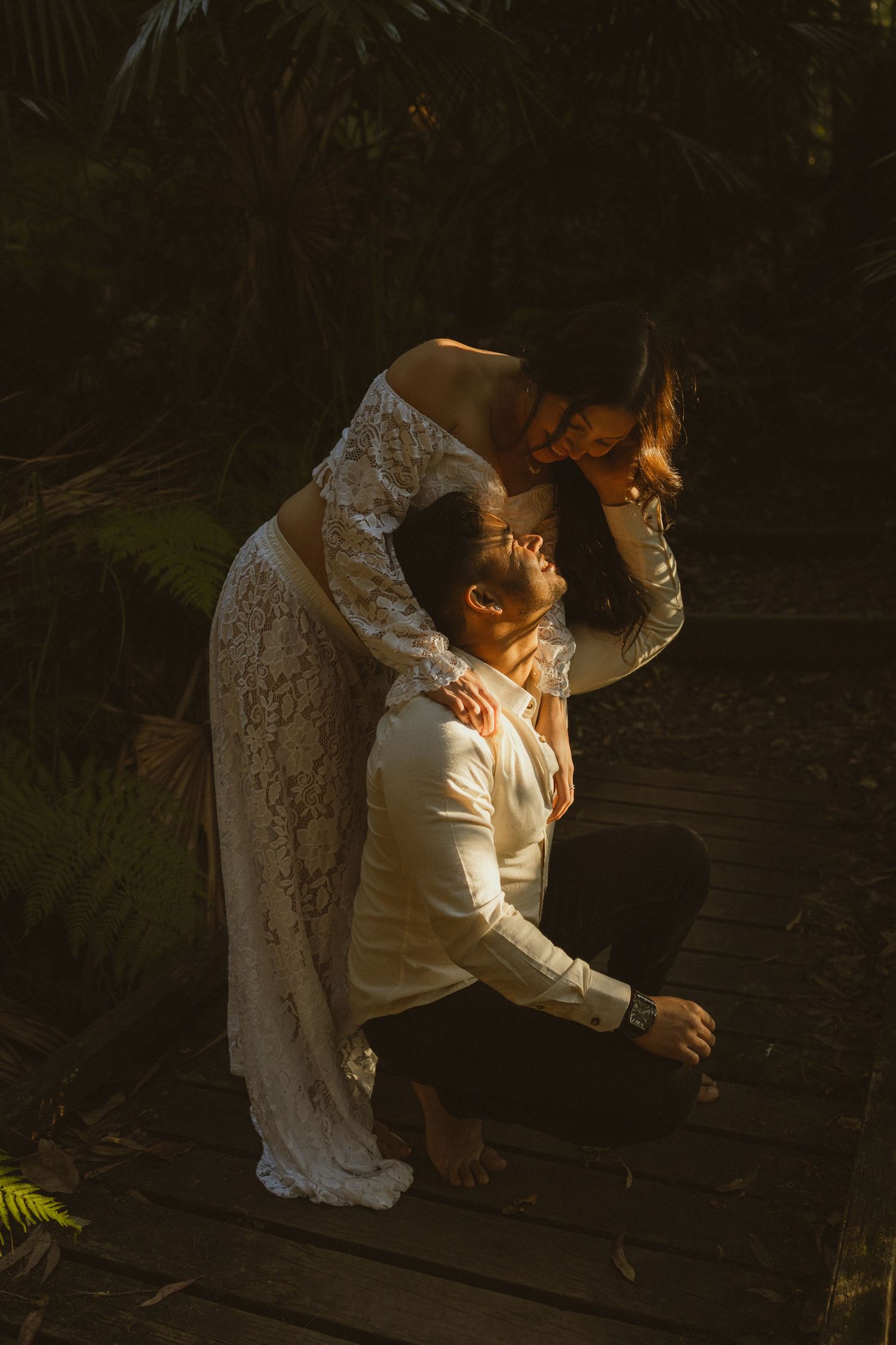 Couple sharing a romantic moment in a sunlit garden, bride in lace dress leaning over groom kneeling on wooden deck.