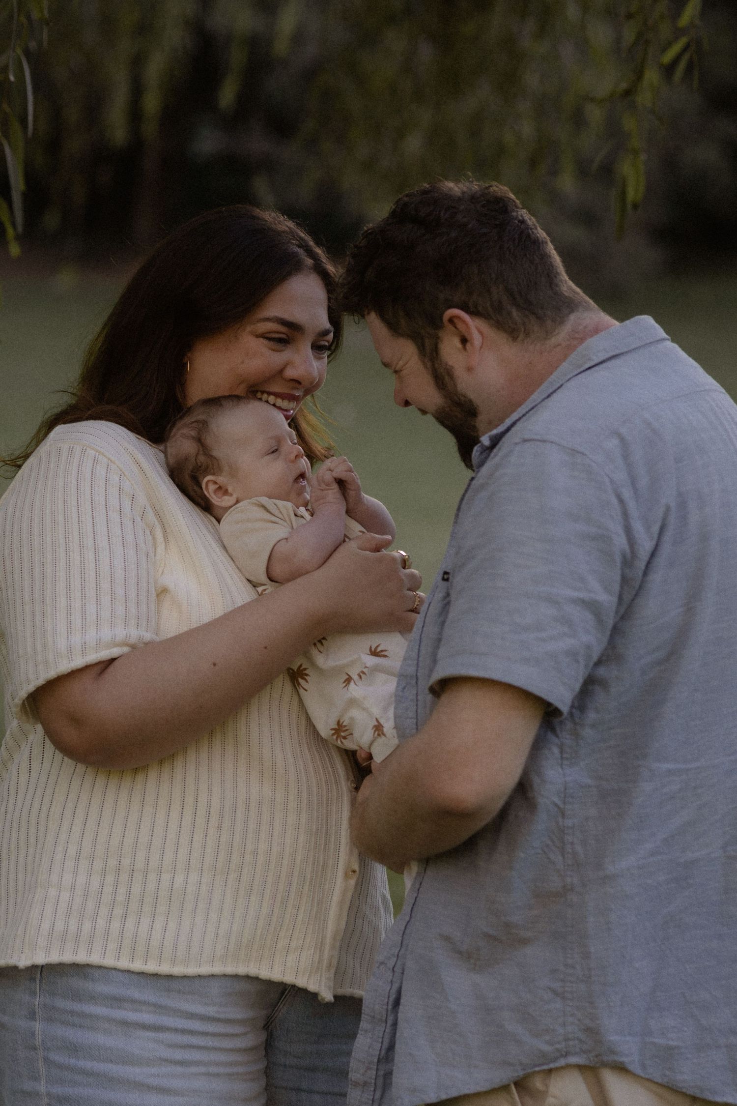 A couple smiling lovingly at their newborn baby outdoors near green willow trees in soft natural light.