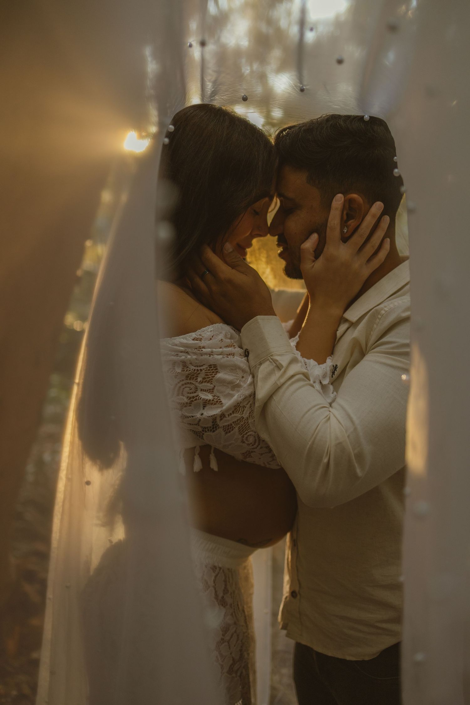 Intimate couple embracing tenderly with foreheads touching, surrounded by warm fairy lights and flowing white fabric.