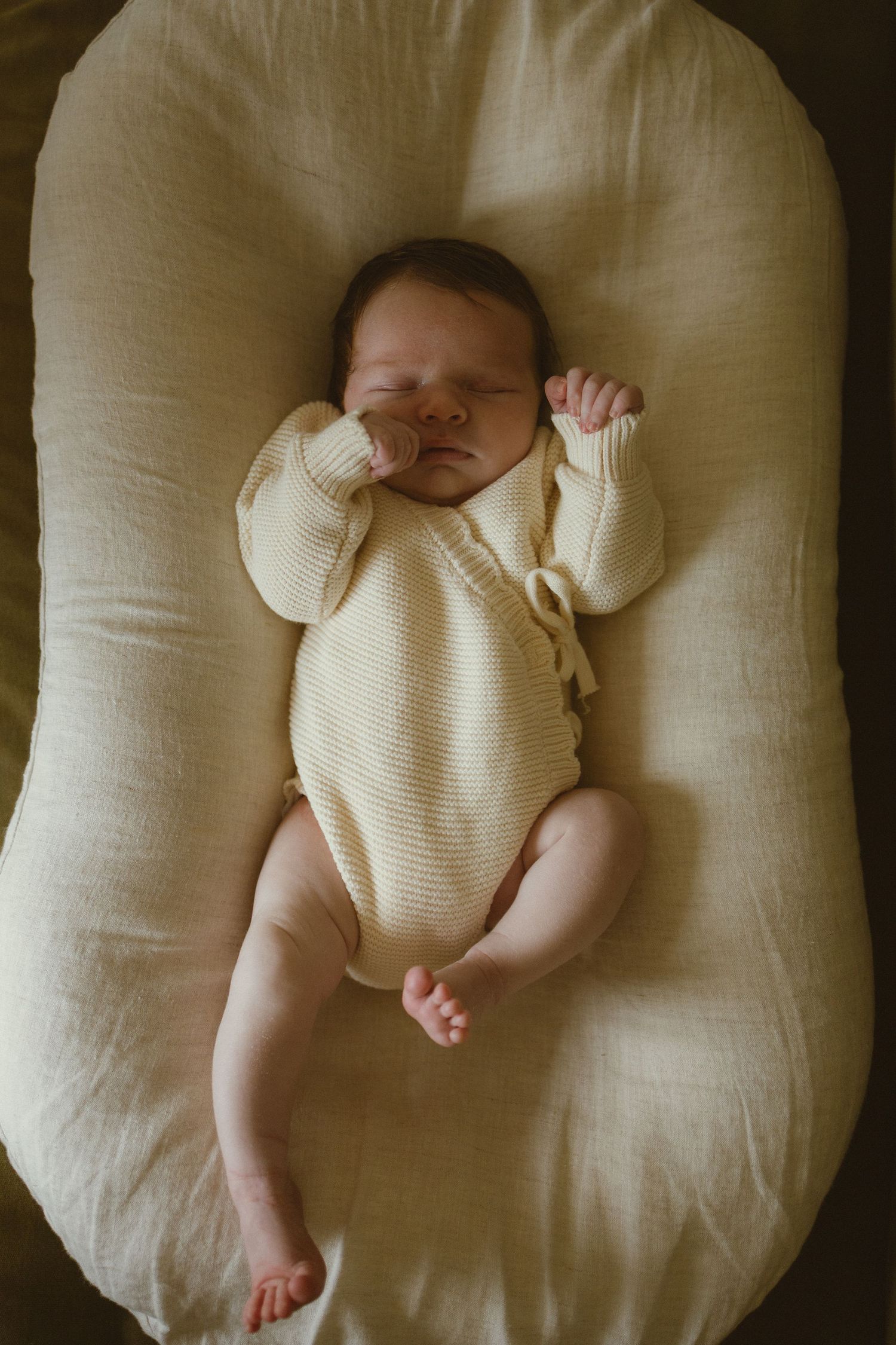 Newborn baby sleeping peacefully in a cream knit romper on a soft ivory lounger pillow.