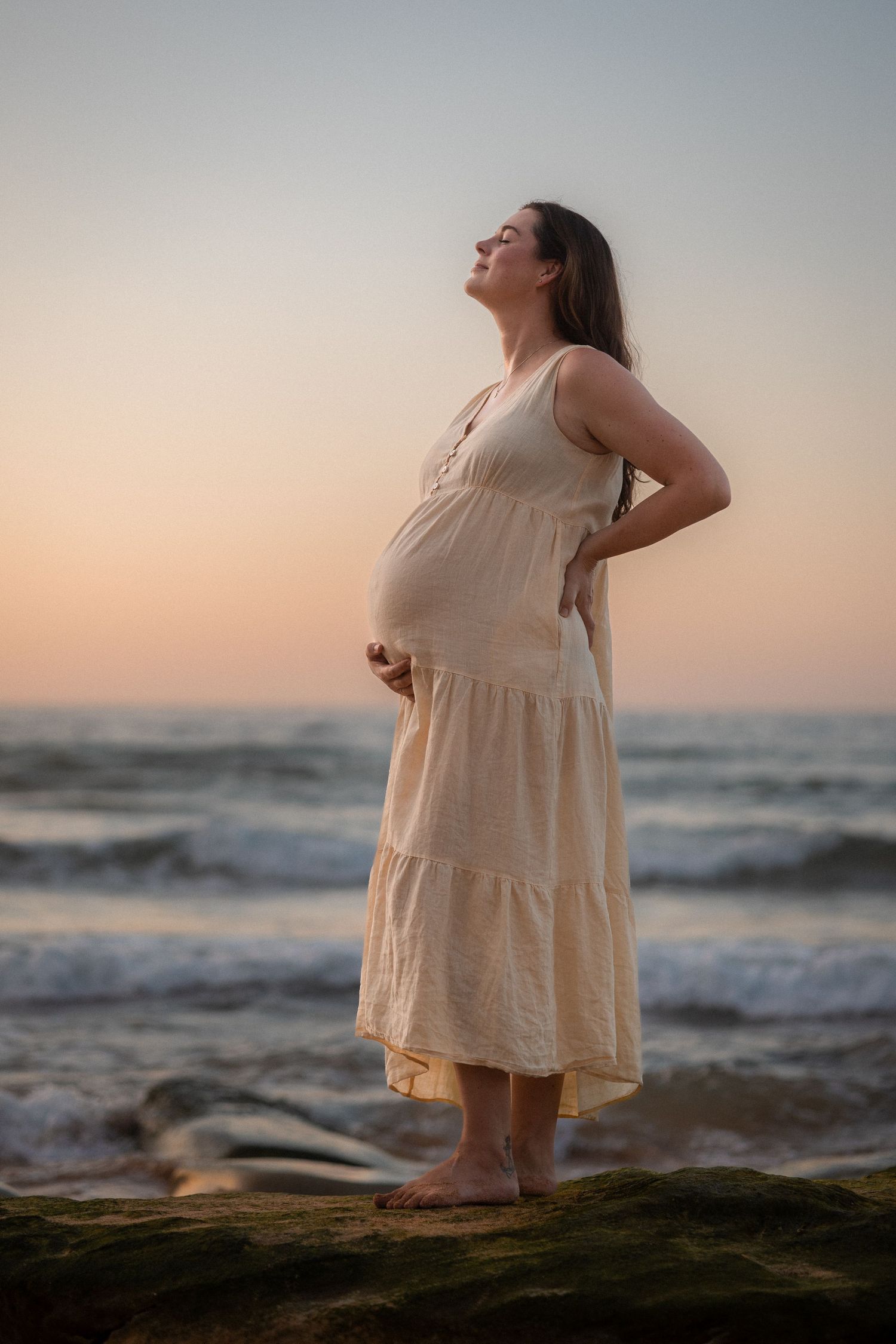Expectant mother in a flowing white dress stands barefoot at the beach during sunset, cradling her baby bump.