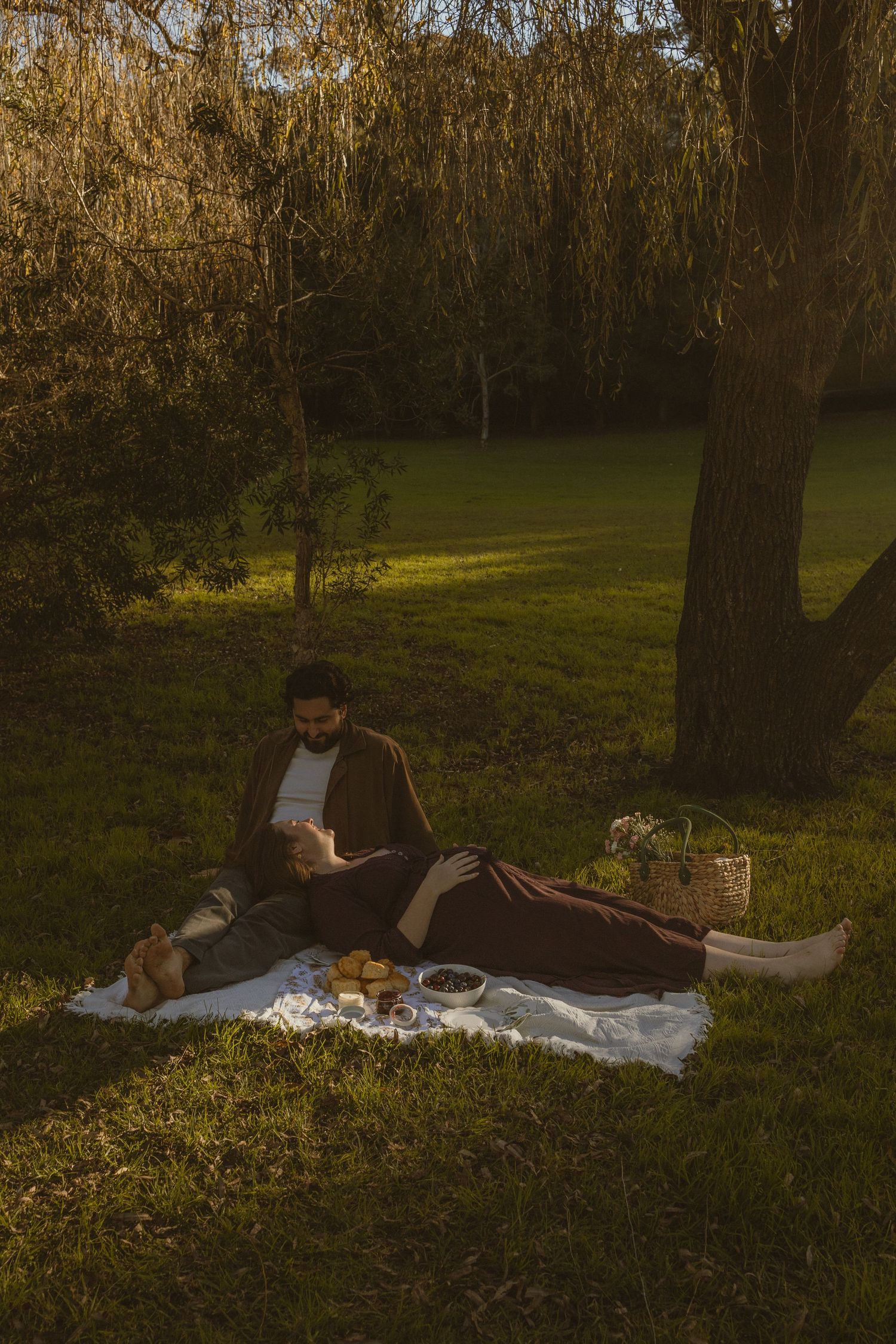 Couple enjoying a romantic picnic on a blanket under a willow tree in golden hour sunlight.