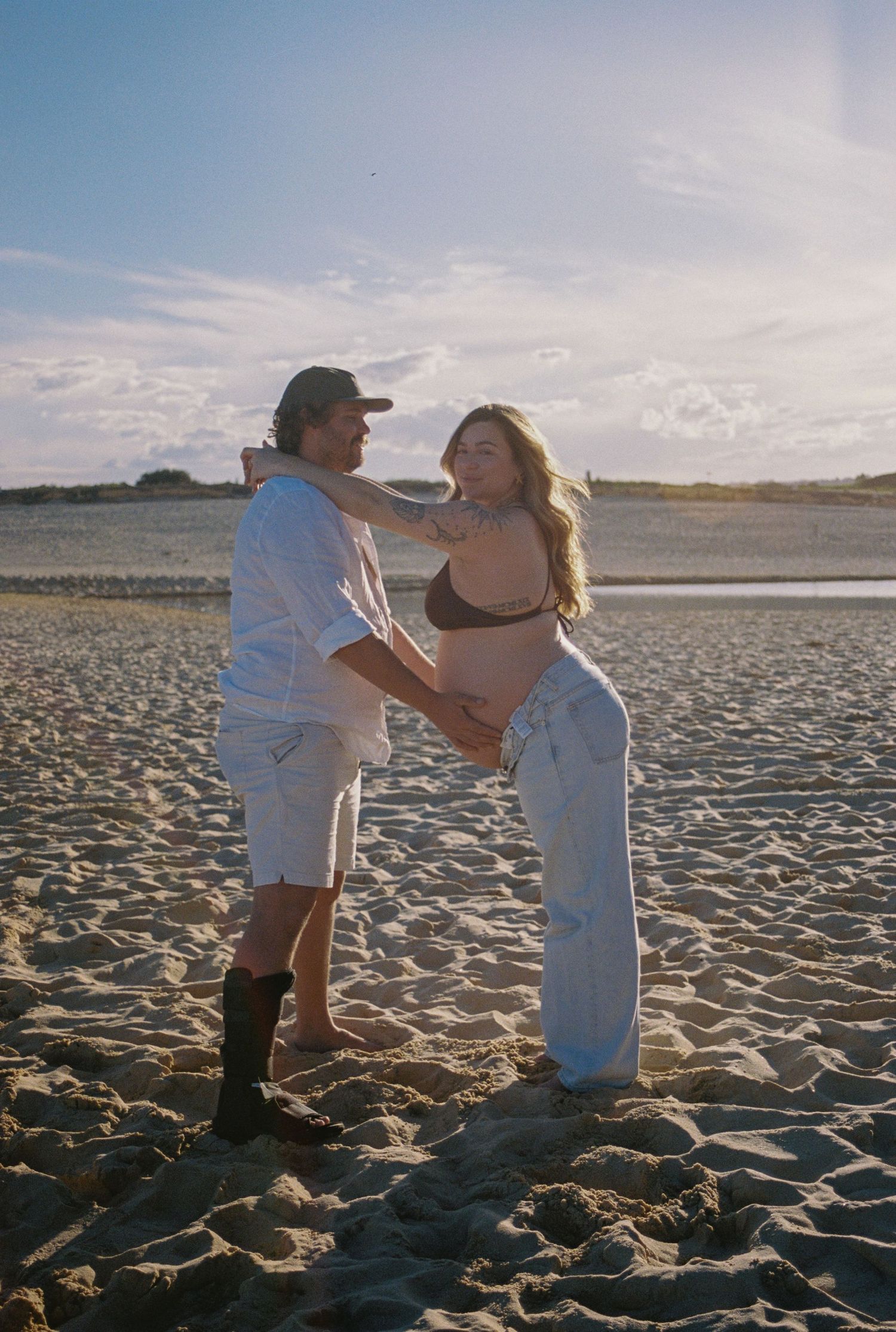 Couple embracing on sandy beach at golden hour, woman showing baby bump in bikini top and jeans.