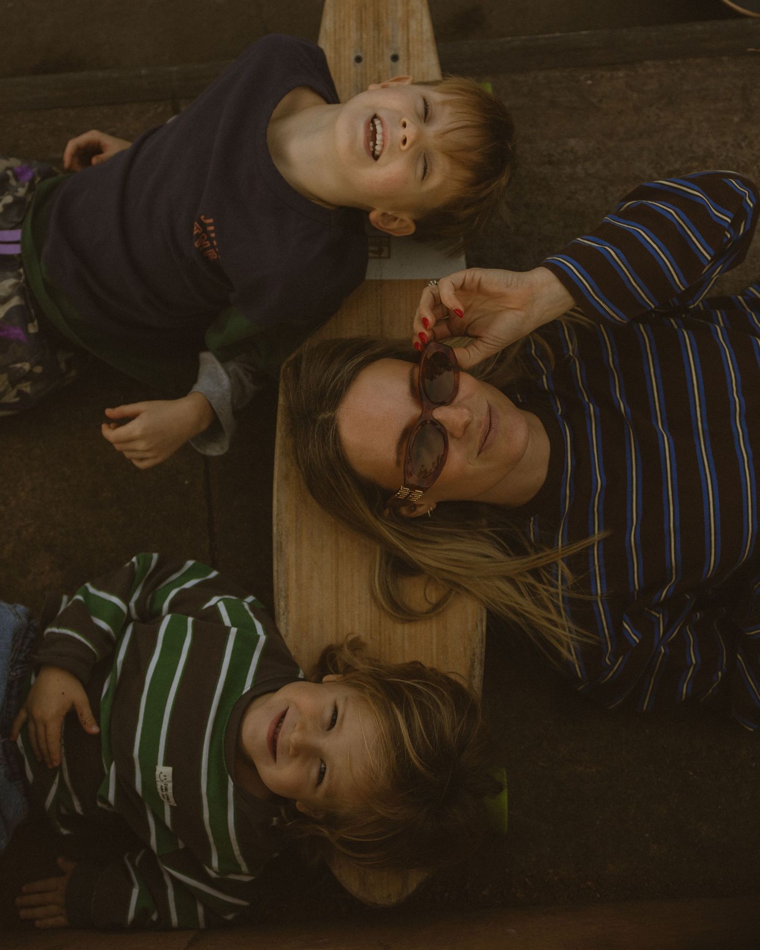 A woman wearing striped shirt and sunglasses lies on wood with two laughing children outdoors.