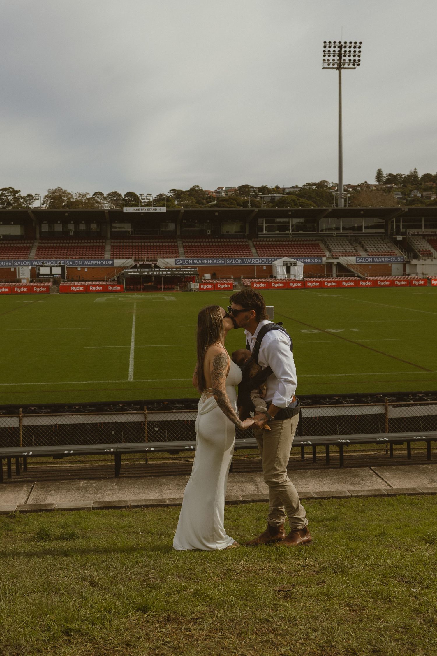 Couple in wedding attire sharing a romantic moment at a sports stadium with green field backdrop.