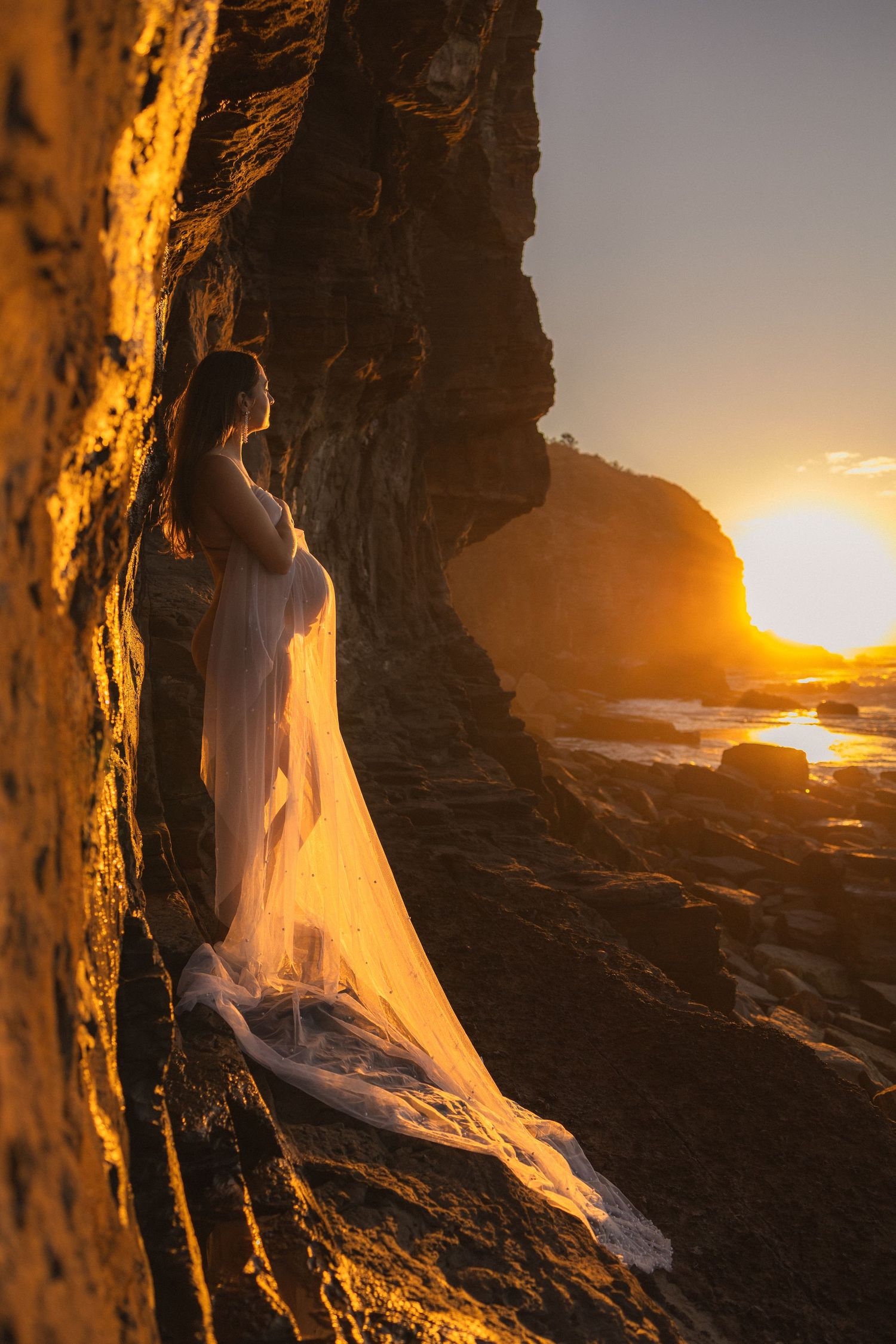 Pregnant woman in flowing white gown standing between coastal rocks at golden sunset.