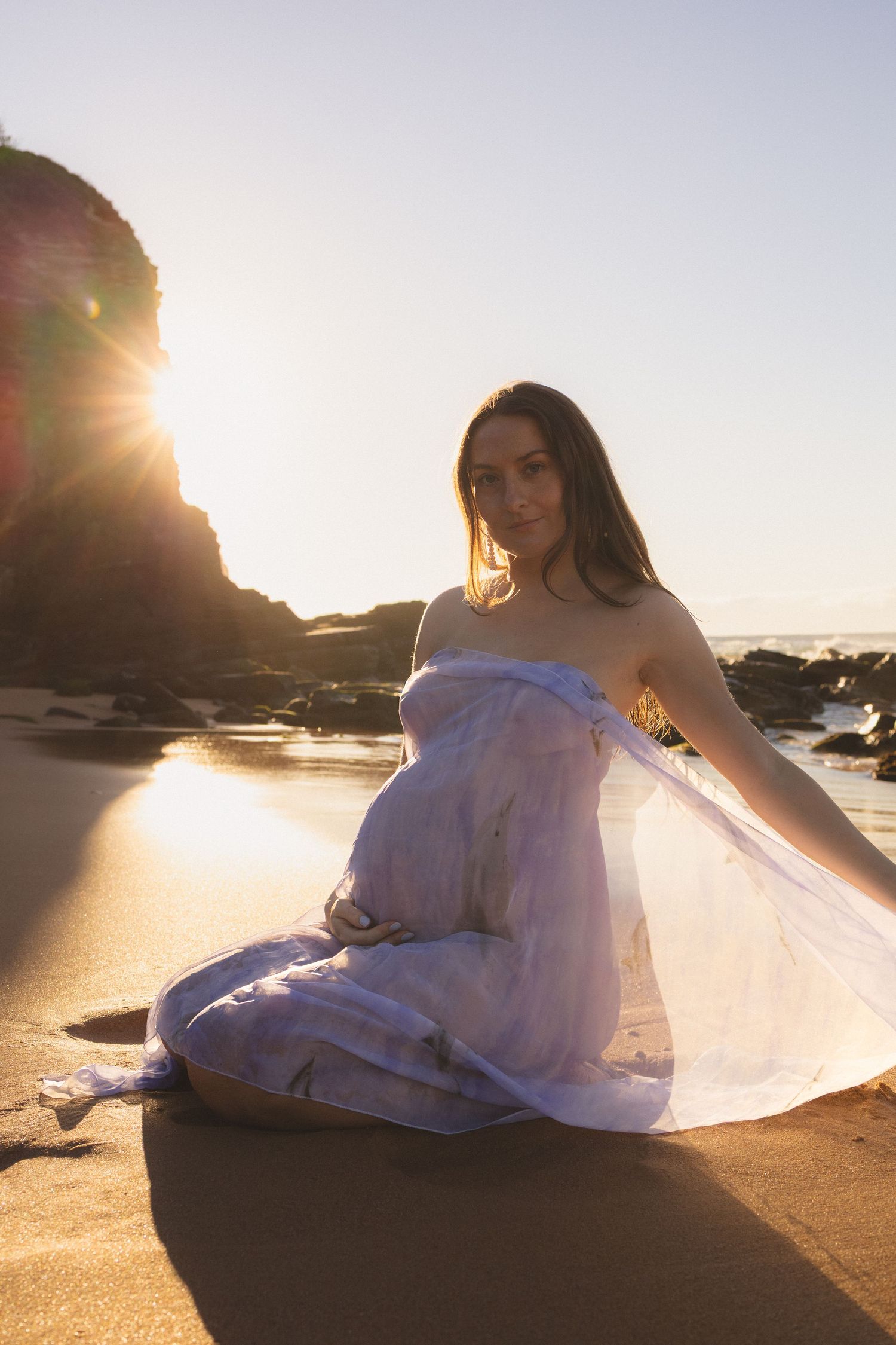 Pregnant woman in flowing white dress sitting on sandy beach glowing in warm sunset light.
