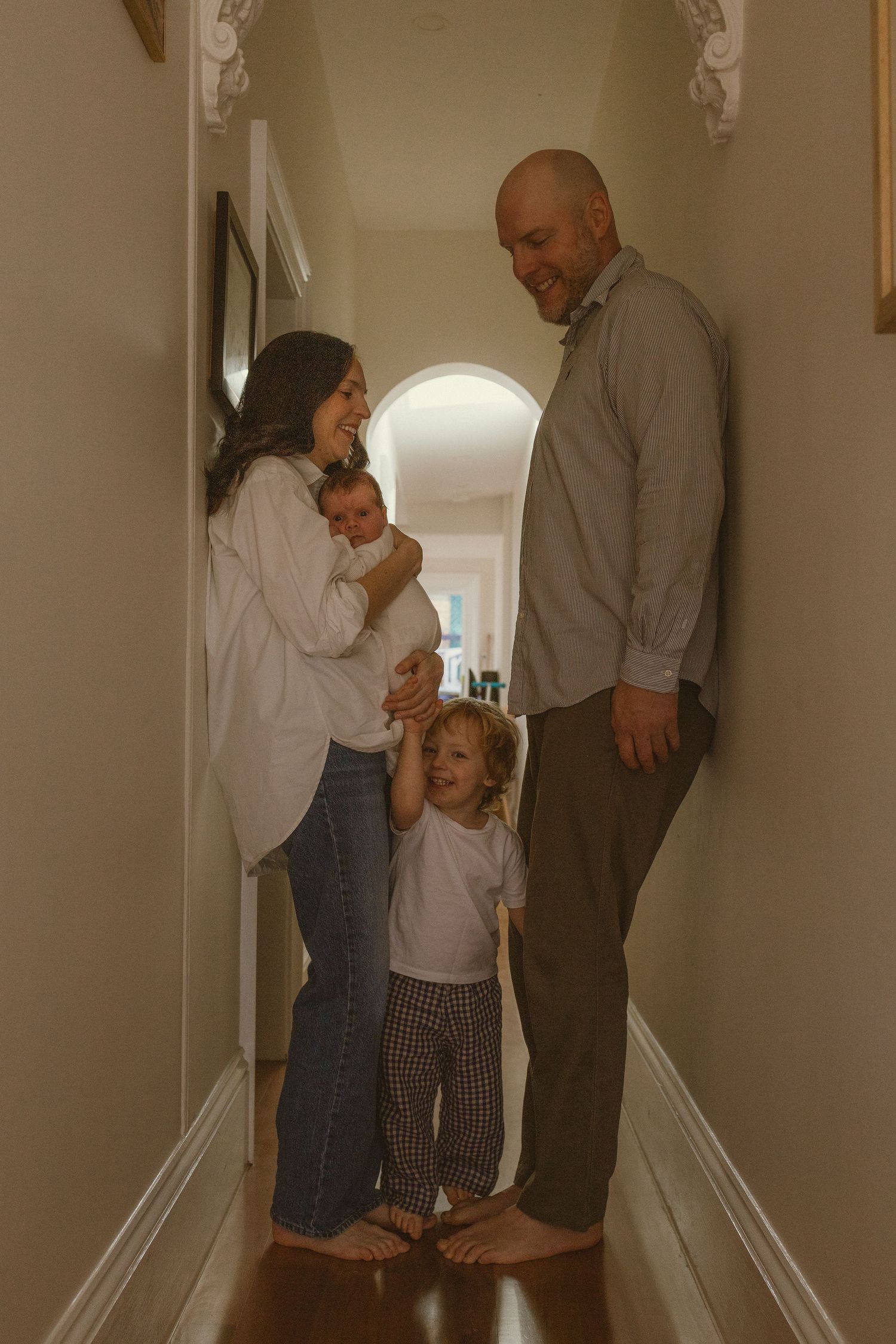 A family of four poses in a hallway, with a mother holding a newborn, a toddler standing between the parents.