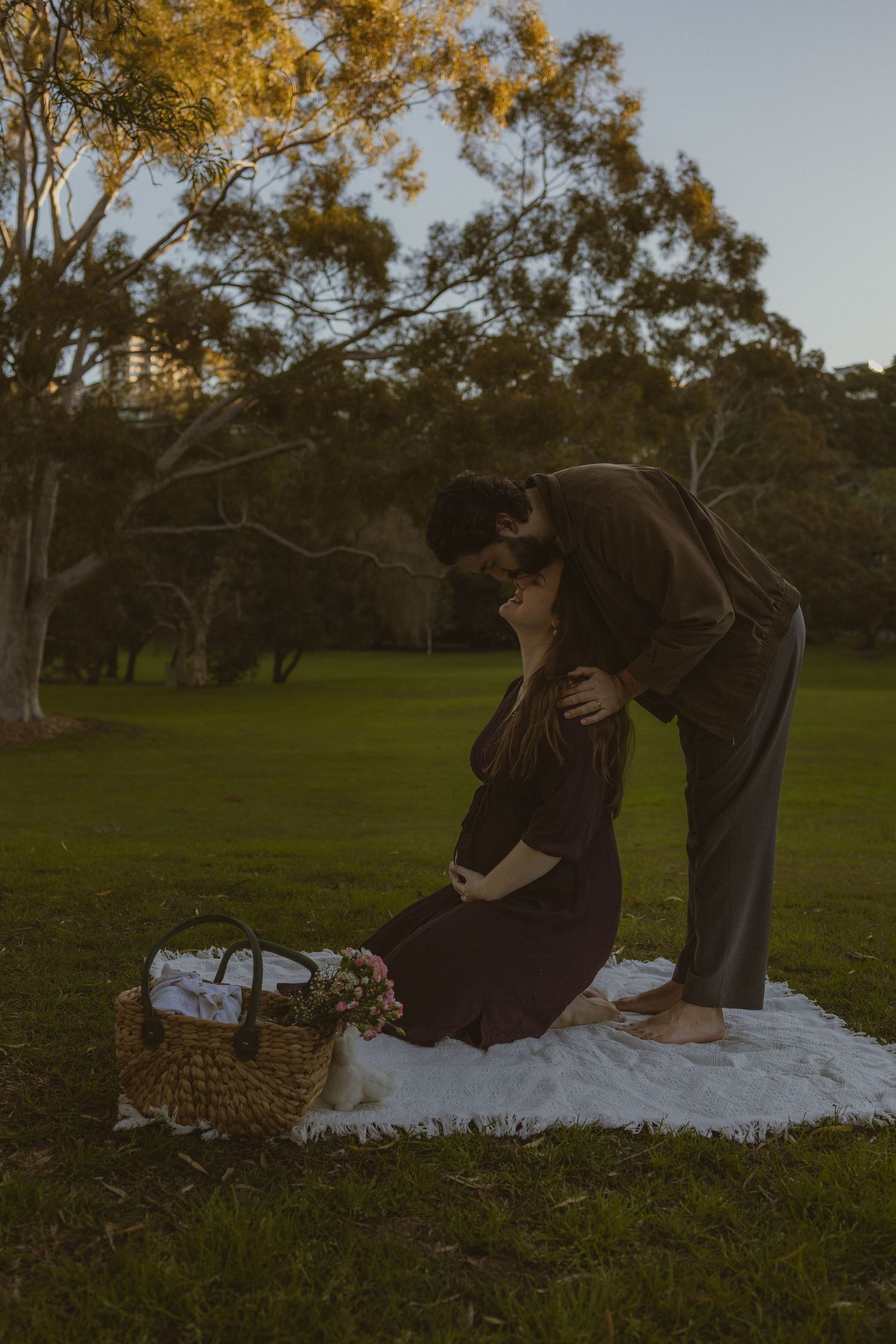 Couple sharing a tender moment at sunset in a park, pregnant woman sitting on blanket with picnic basket nearby.