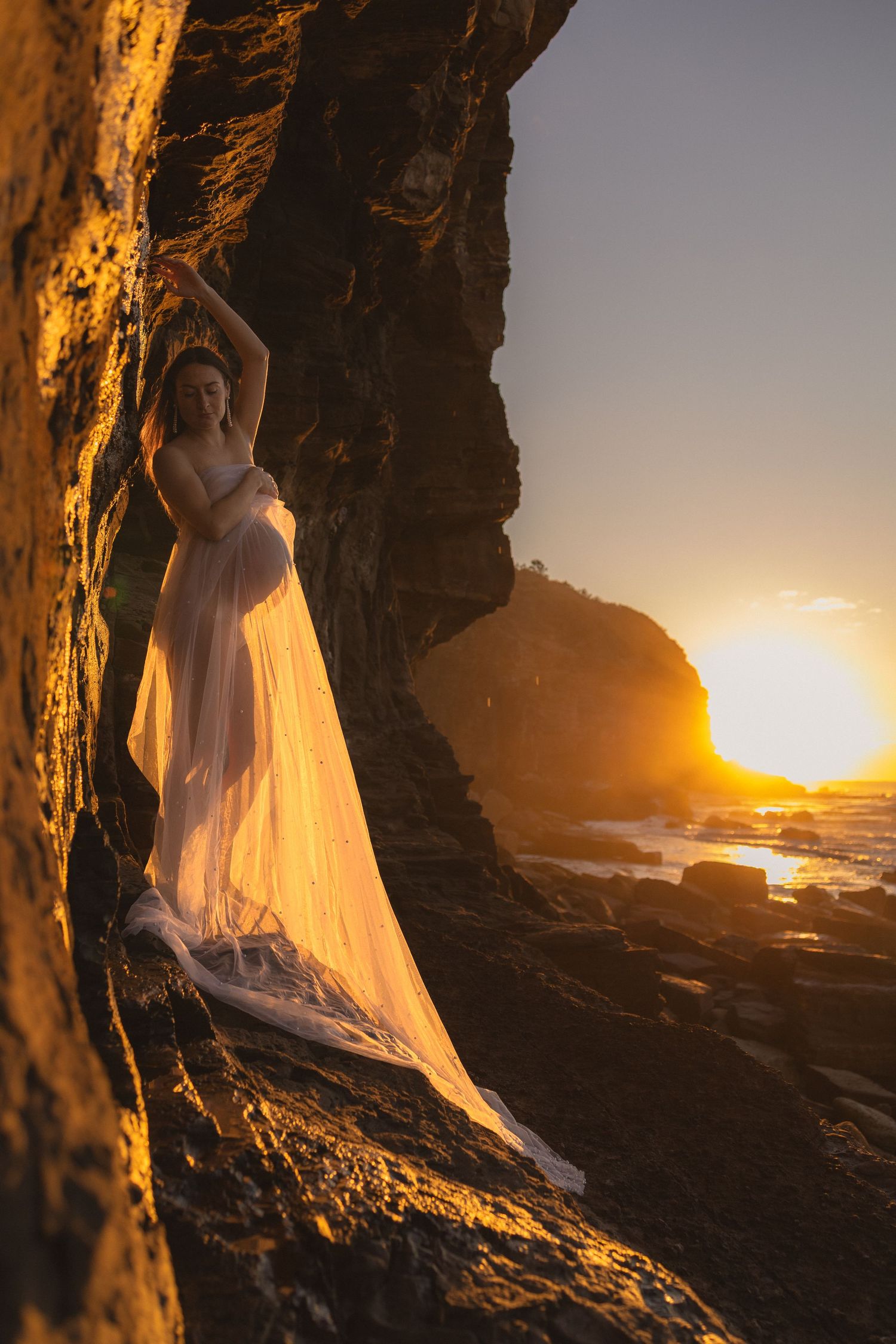 Expectant mother in elegant white maternity gown posing gracefully on rocky beach at sunset.