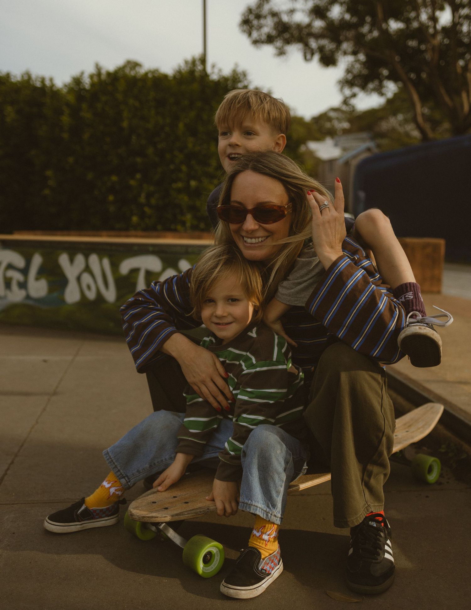 A smiling woman in sunglasses sits at a skate park with two young boys on a skateboard, all wearing striped tops.