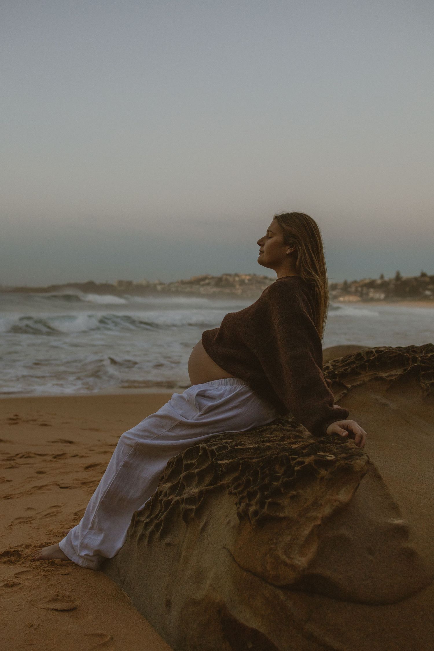 Pregnant woman in white pants sits on beach rock at dusk, gazing at ocean waves with coastal town in background.