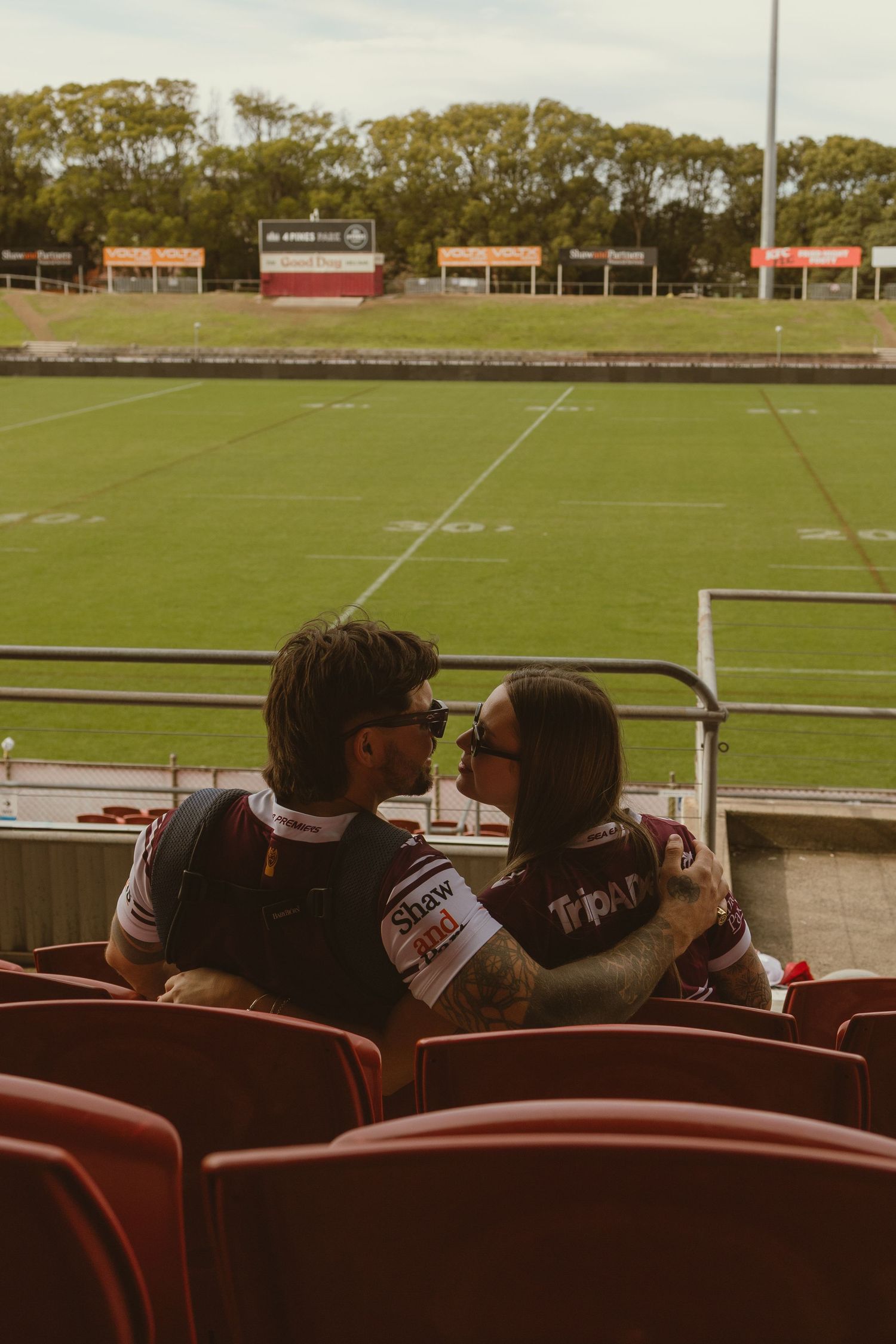 Couple sharing a kiss in stadium seats wearing matching jerseys, with a green rugby field in the background.