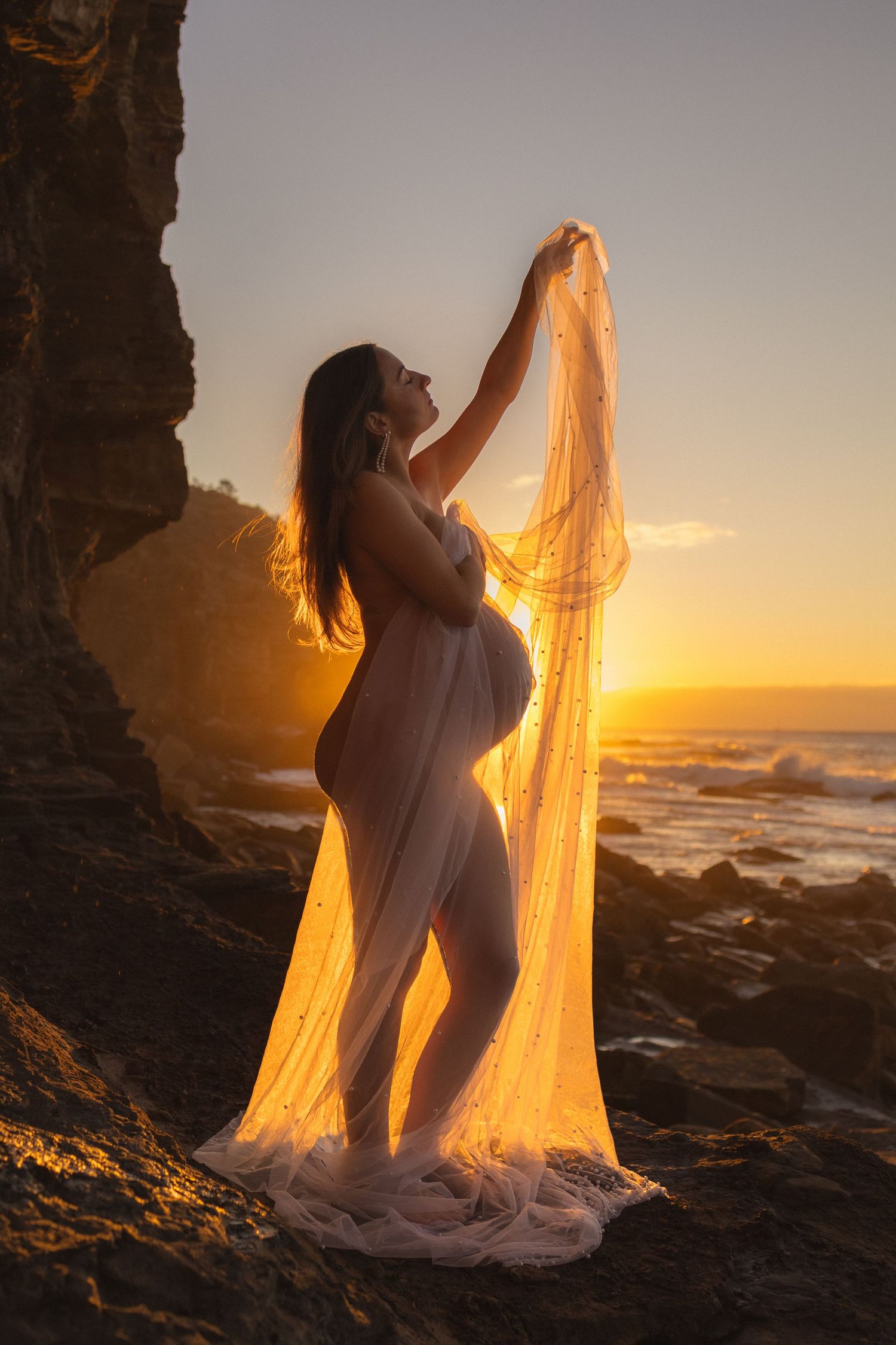Pregnant woman in sheer white fabric poses on rocky beach at golden sunset with ocean waves.