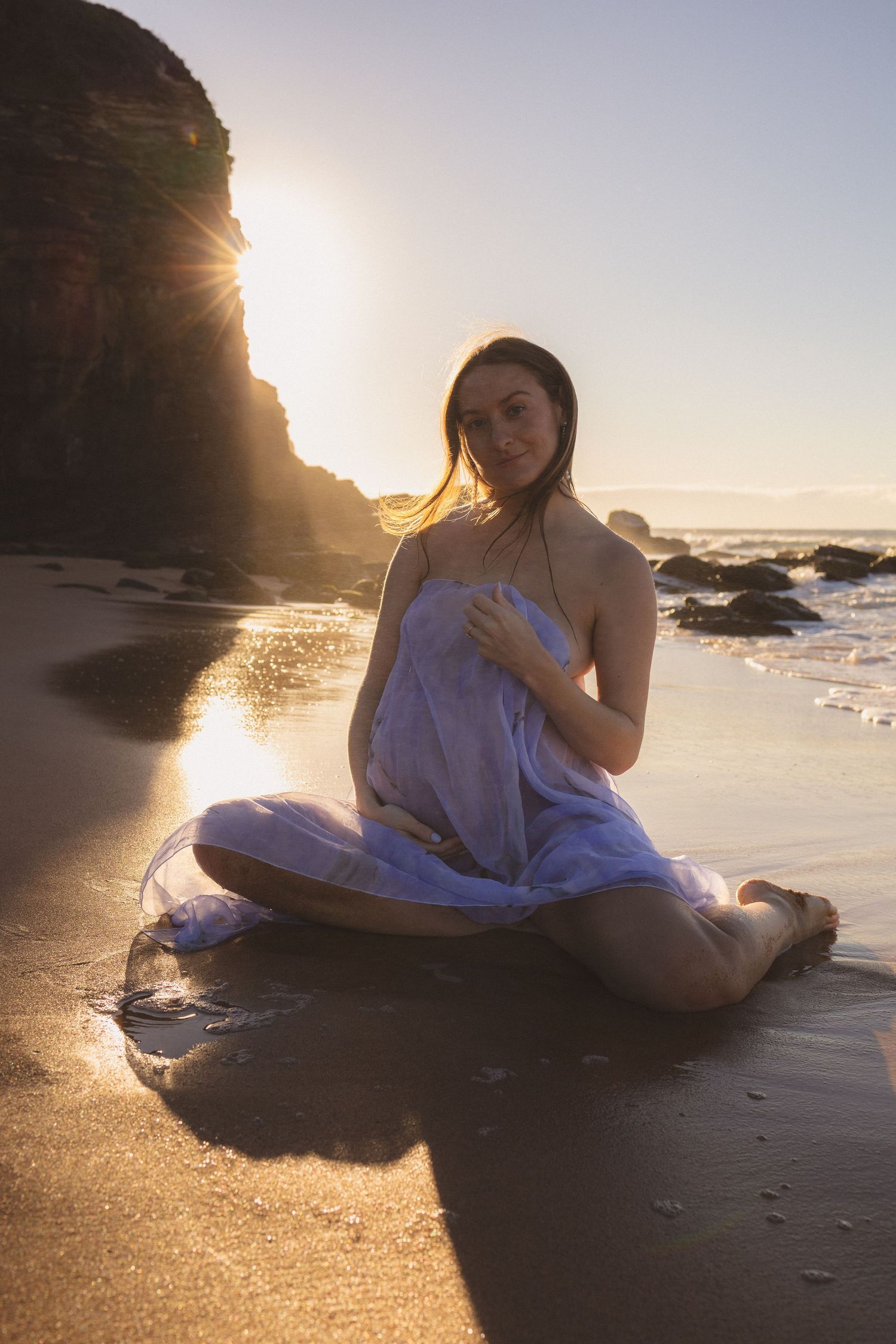 Pregnant woman in sheer wrap sits cross-legged on wet sandy beach during golden sunset.