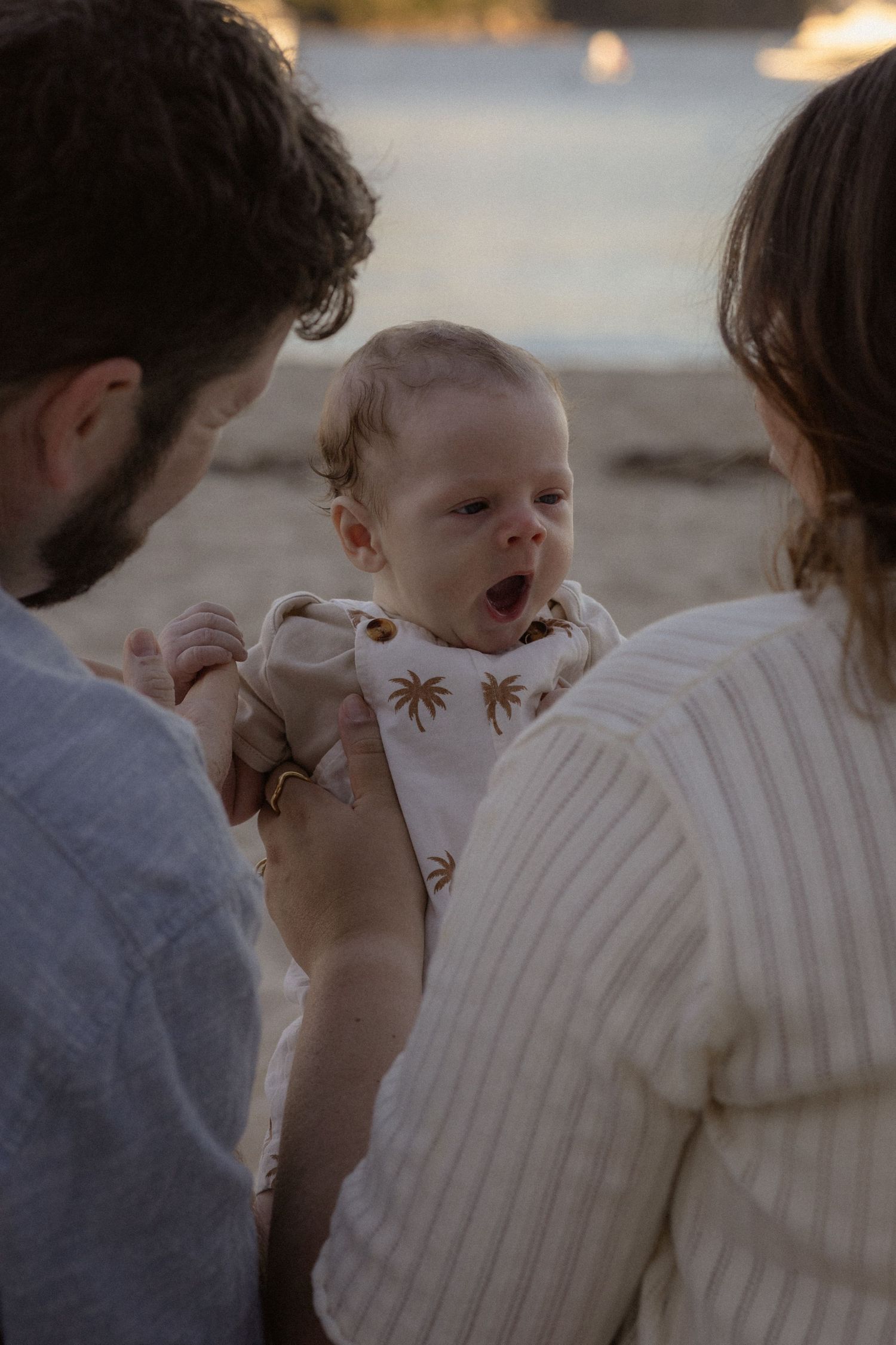 Parents holding a yawning baby on a sandy beach at golden hour, with water in the background.