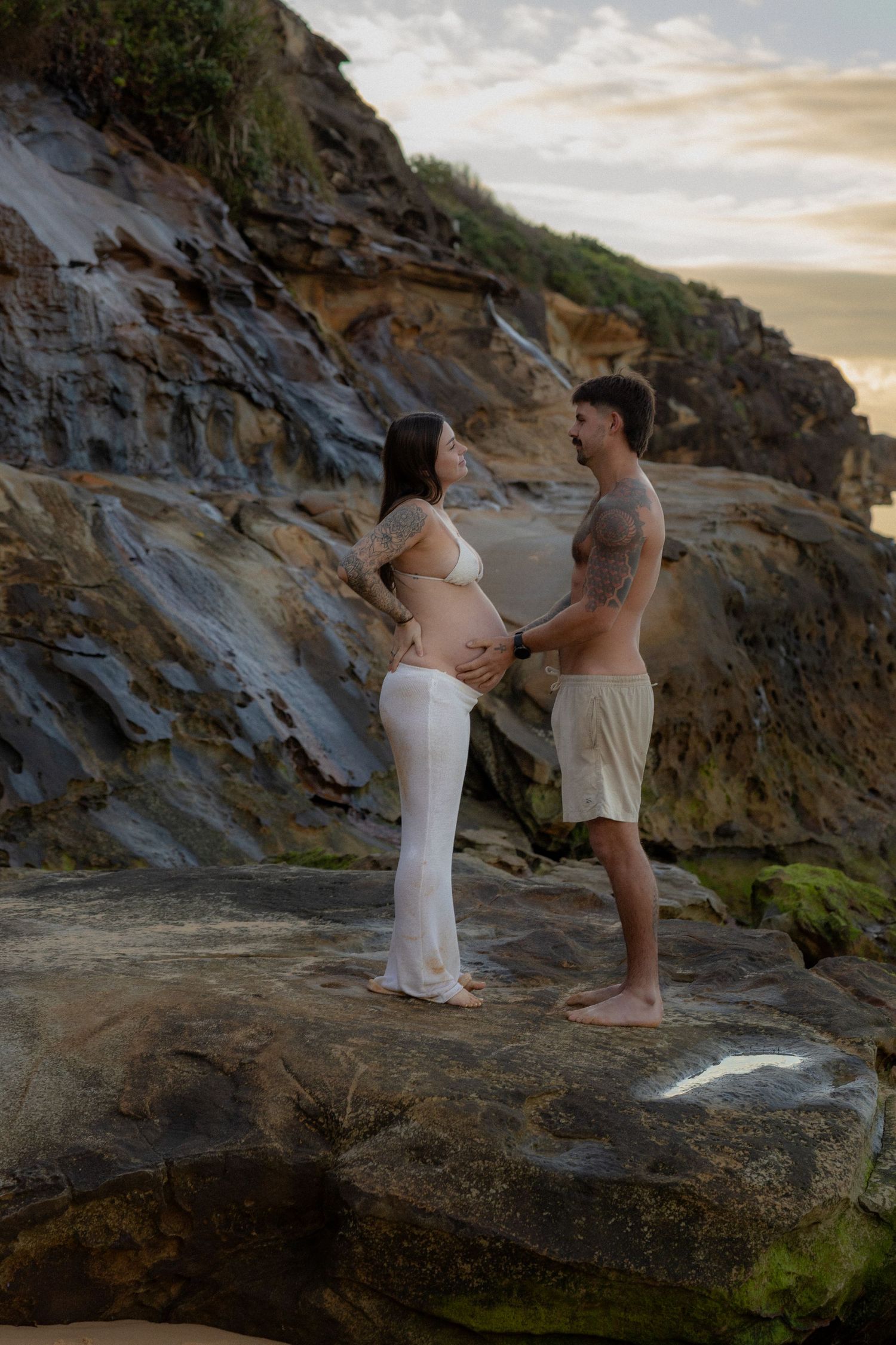 Expecting couple standing on coastal rocks at sunset, man holds pregnant womans belly near cliff face.