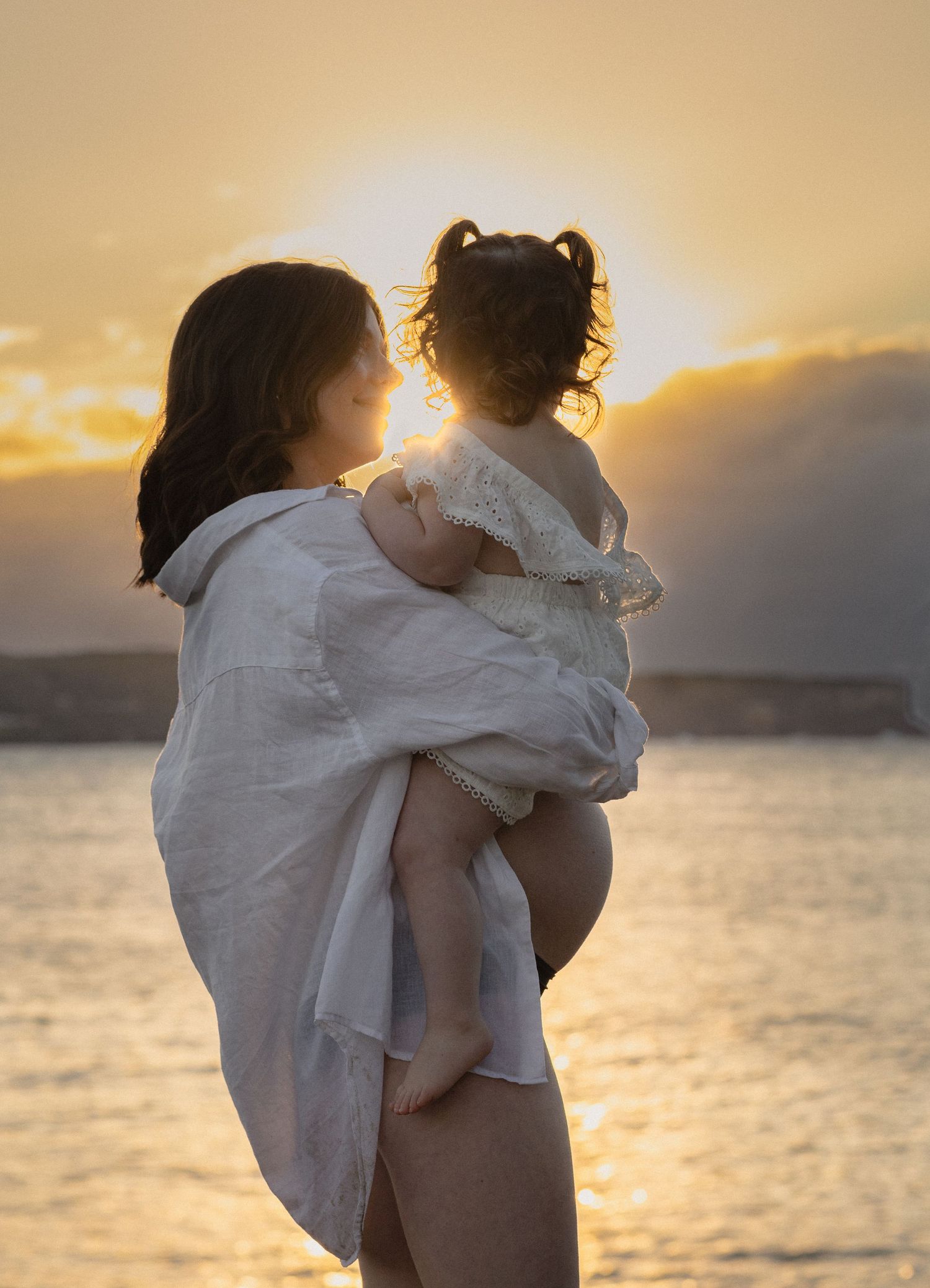 Mother holding toddler at golden sunset by the water, both dressed in white, sharing a tender moment.