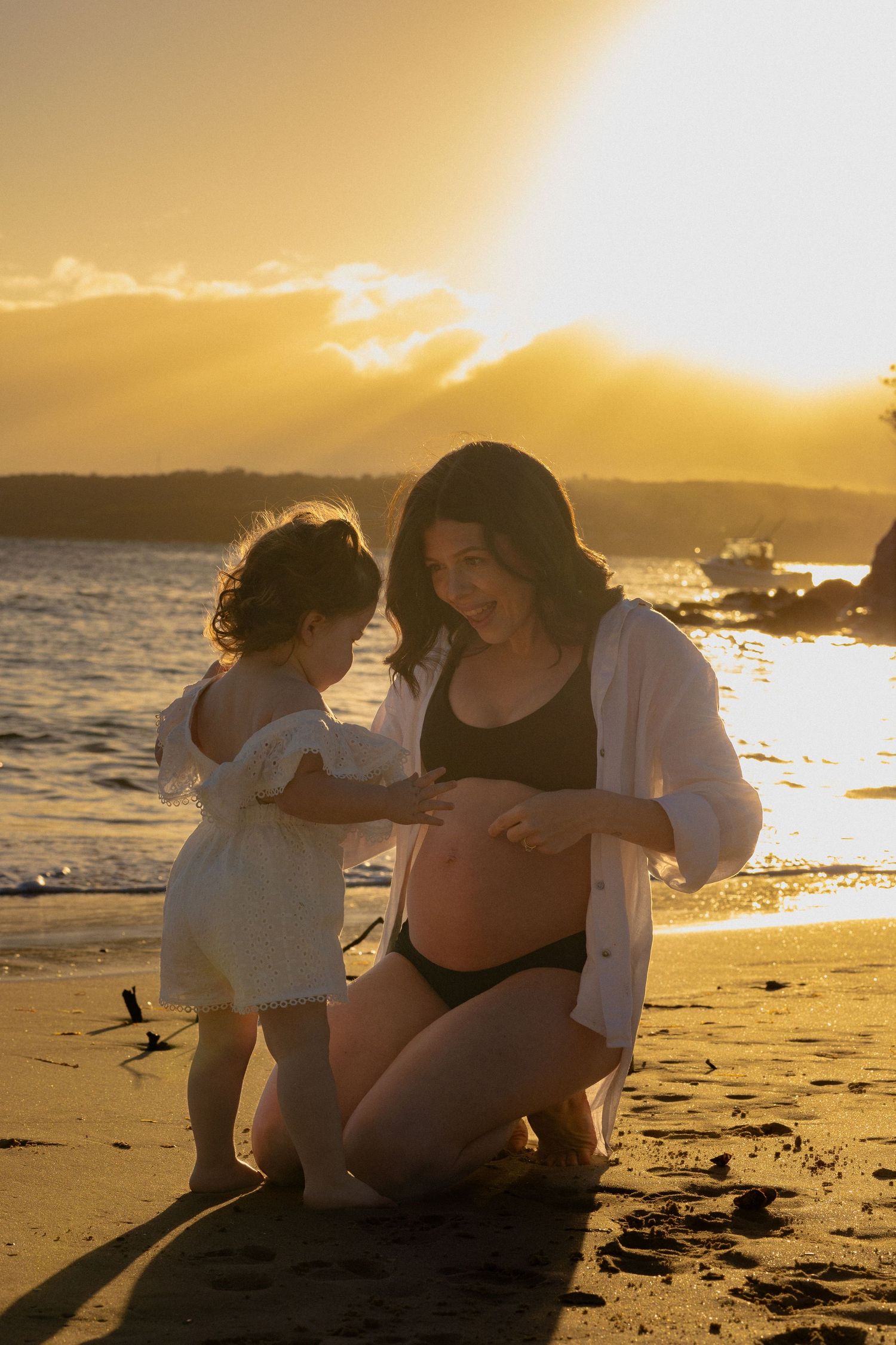 Pregnant woman kneeling on beach at sunset with toddler touching her baby bump, golden light reflecting on ocean.