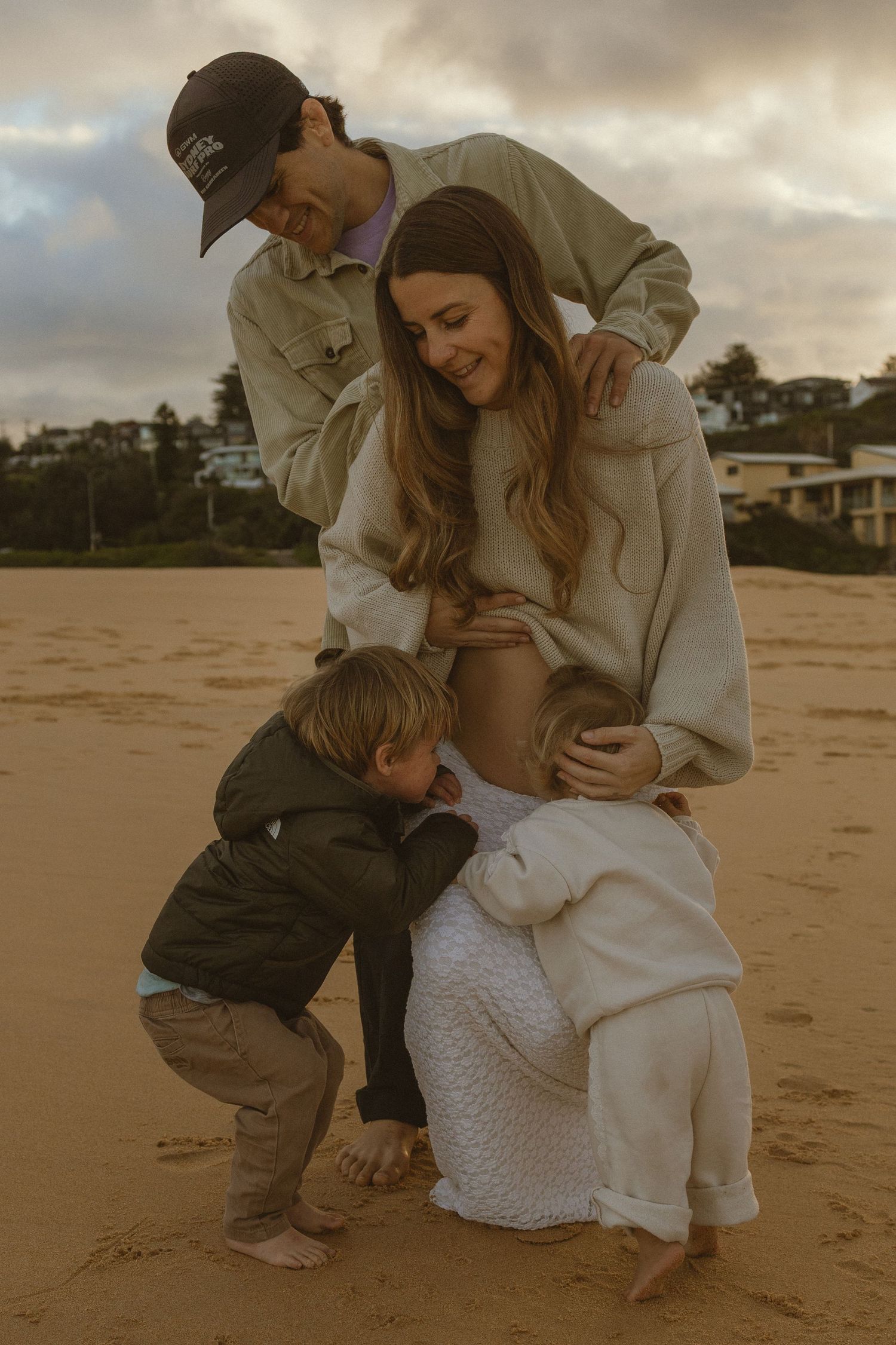 Family maternity photo on beach, pregnant woman with partner and young child on sandy shore at sunset.